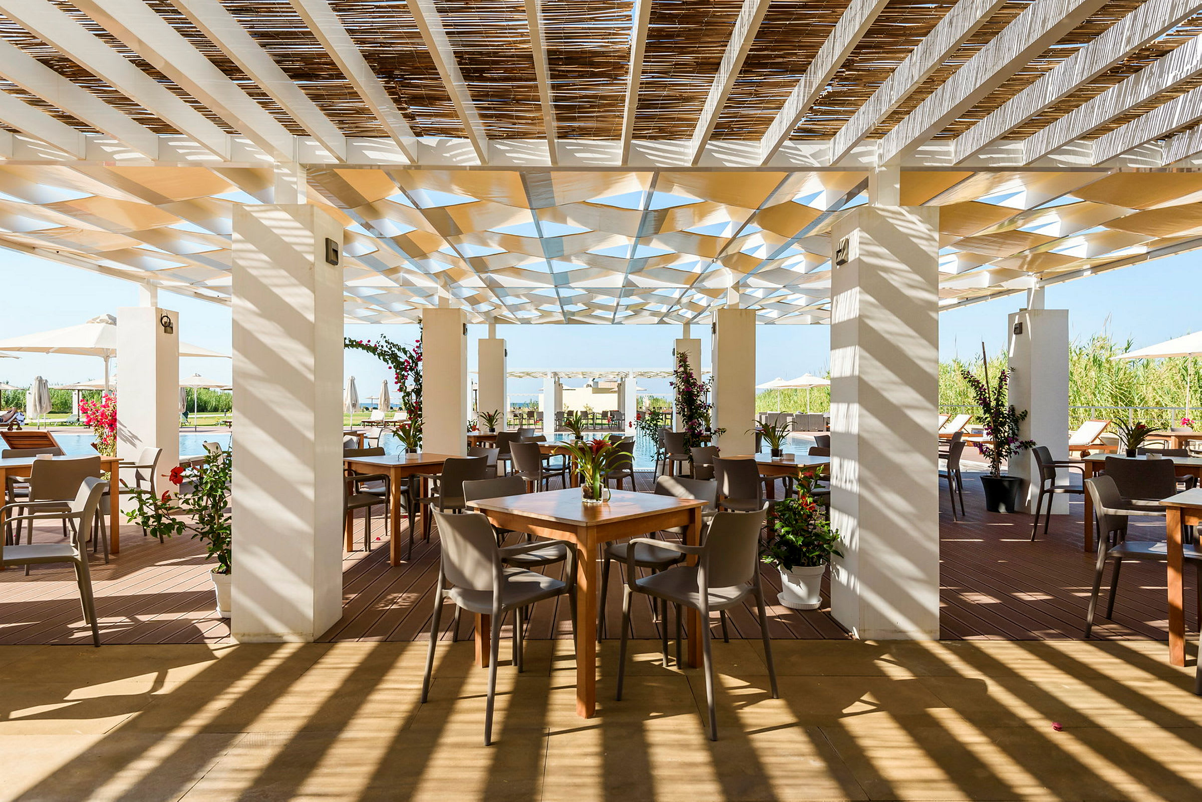 En utomhusrestaurang med bord och stolar under en skuggande pergola, omgiven av grönska. (AI-genererad text)