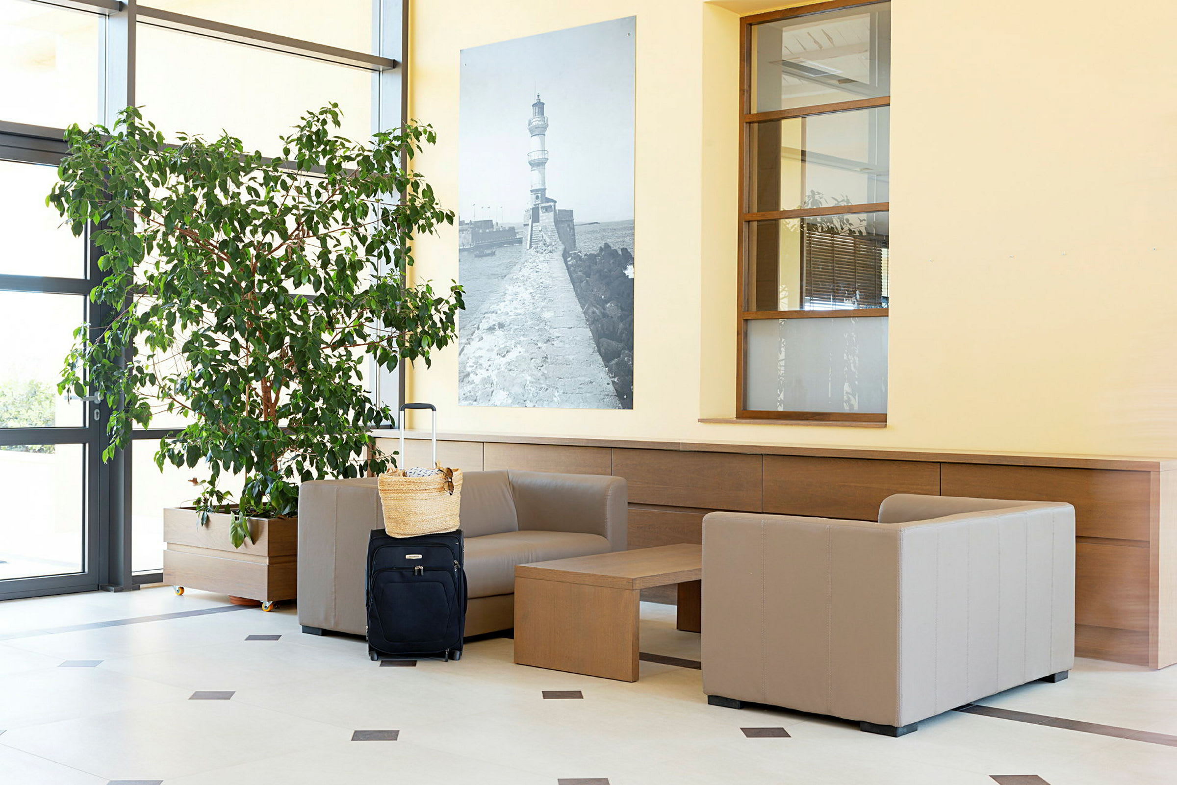 En receptionslounge med beige fåtöljer, ett bord, en stor grön växt samt en svart resväska med en korg ovanpå. (AI-genererad text)