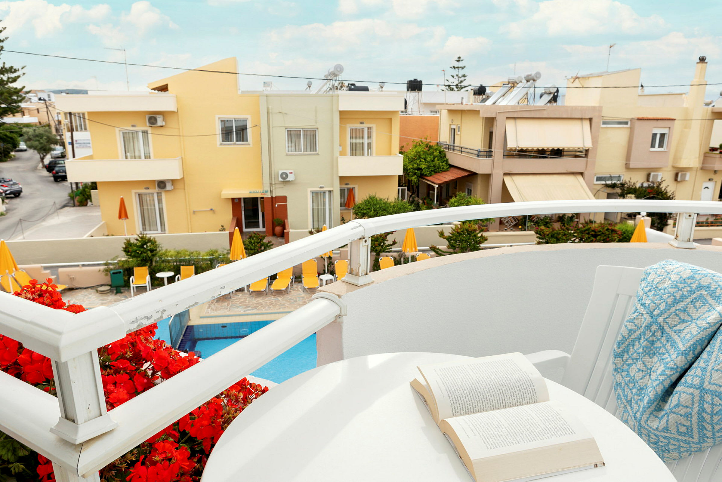 Ett hotellområde med en swimmingpool, omgiven av solstolar och byggnader i bakgrunden, sett från en balkong med bord och stolar. (AI-genererad text)