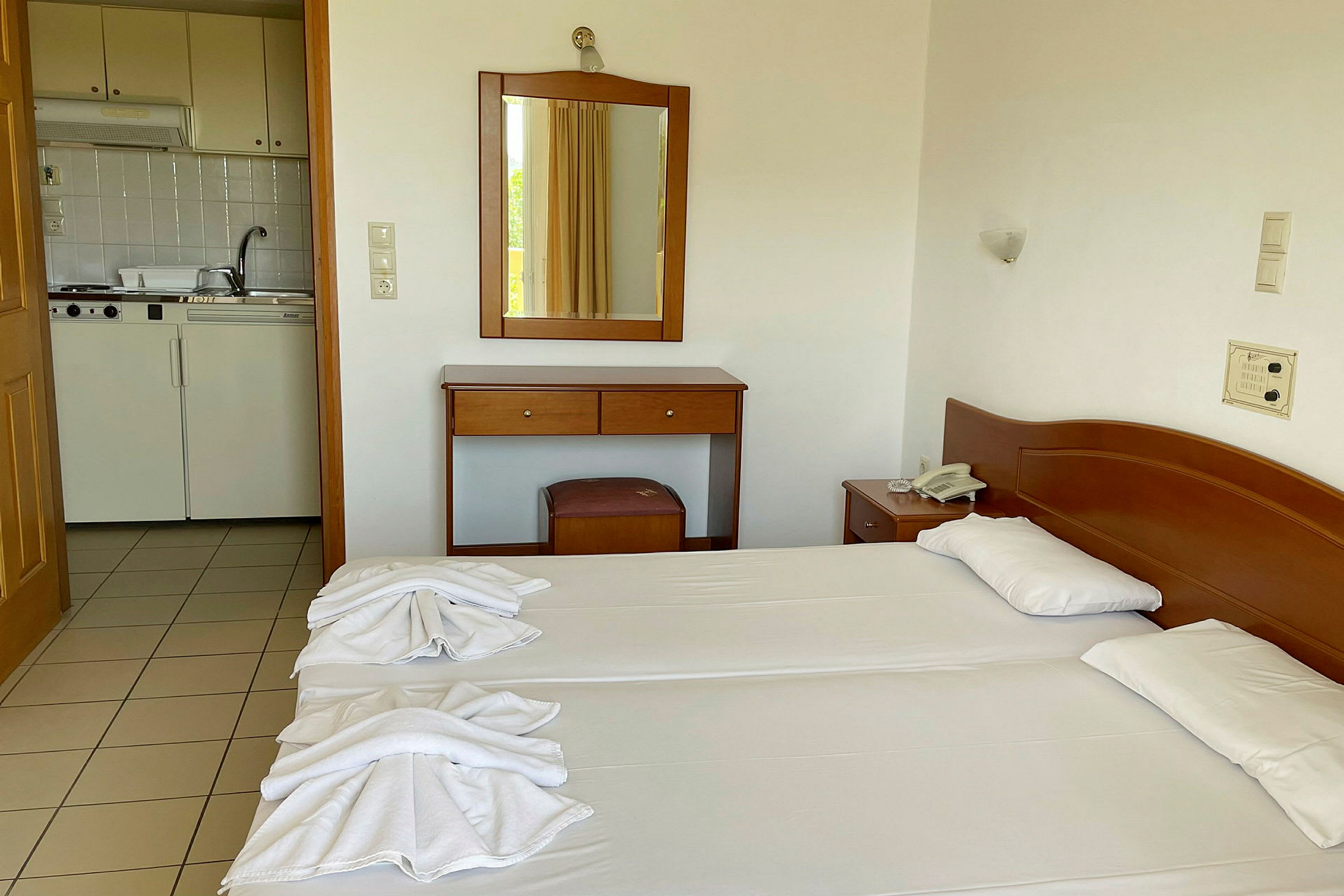 Ett hotellrum med två sängar, ett träskrivbord med spegel och en öppning mot ett kök. (AI-genererad text)