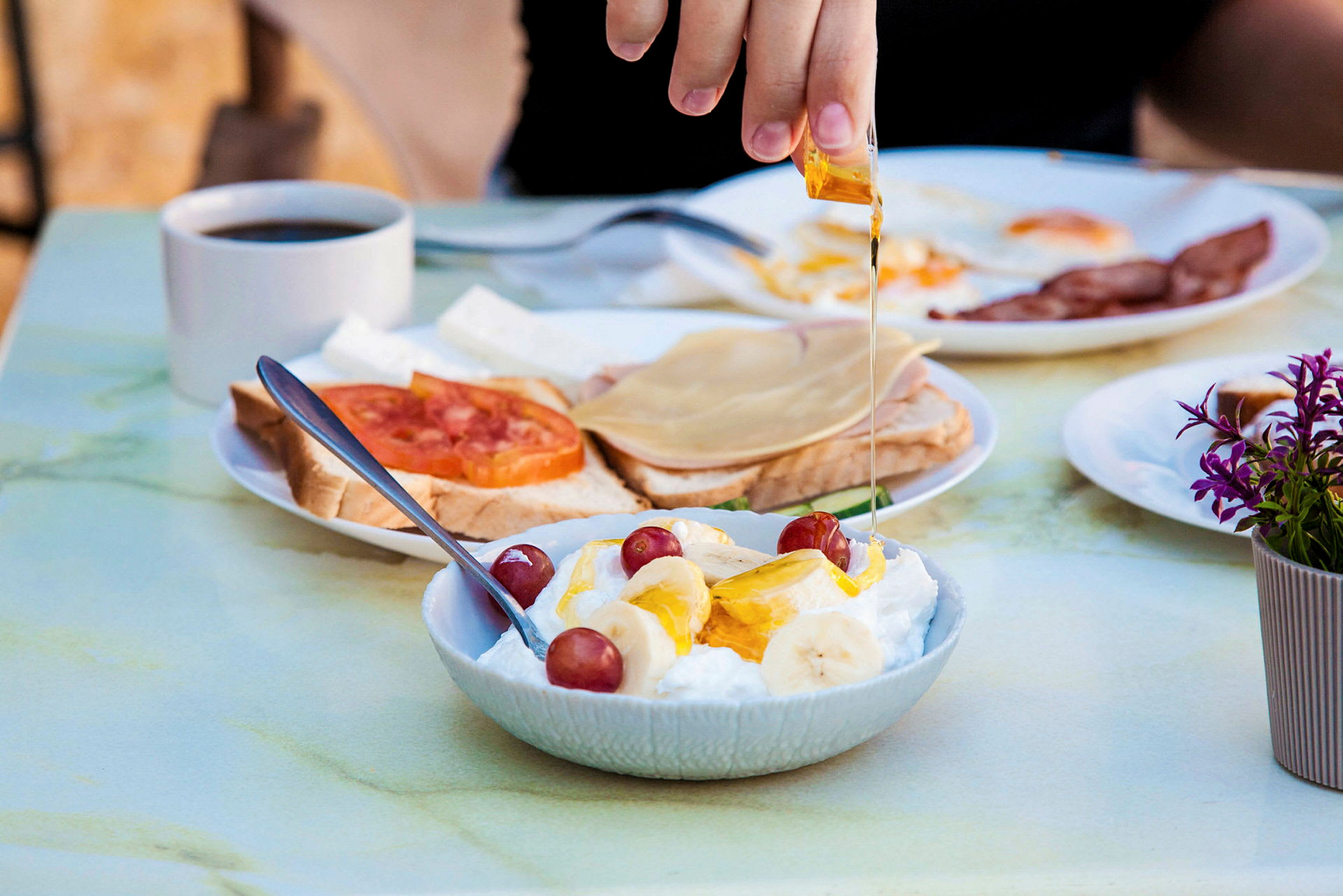 En hand ringlar honung över en skål med yoghurt, banan och vindruvor. I bakgrunden finns en kopp kaffe och tallrikar med bröd och pålägg. (AI-genererad text)