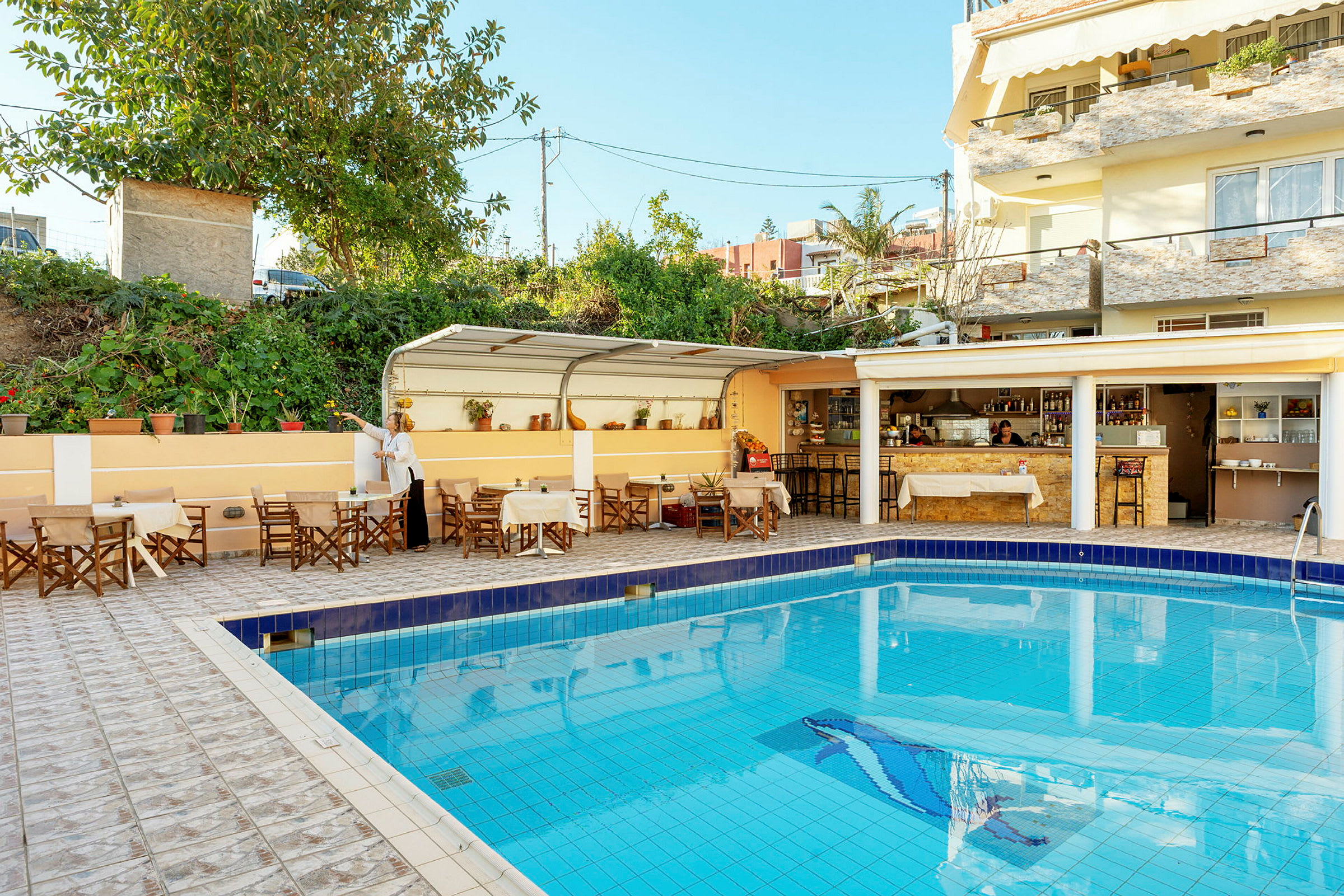Utomhusområde med en pool som omges av bord och stolar. I bakgrunden en täckt café- eller barservering. (AI-genererad text)
