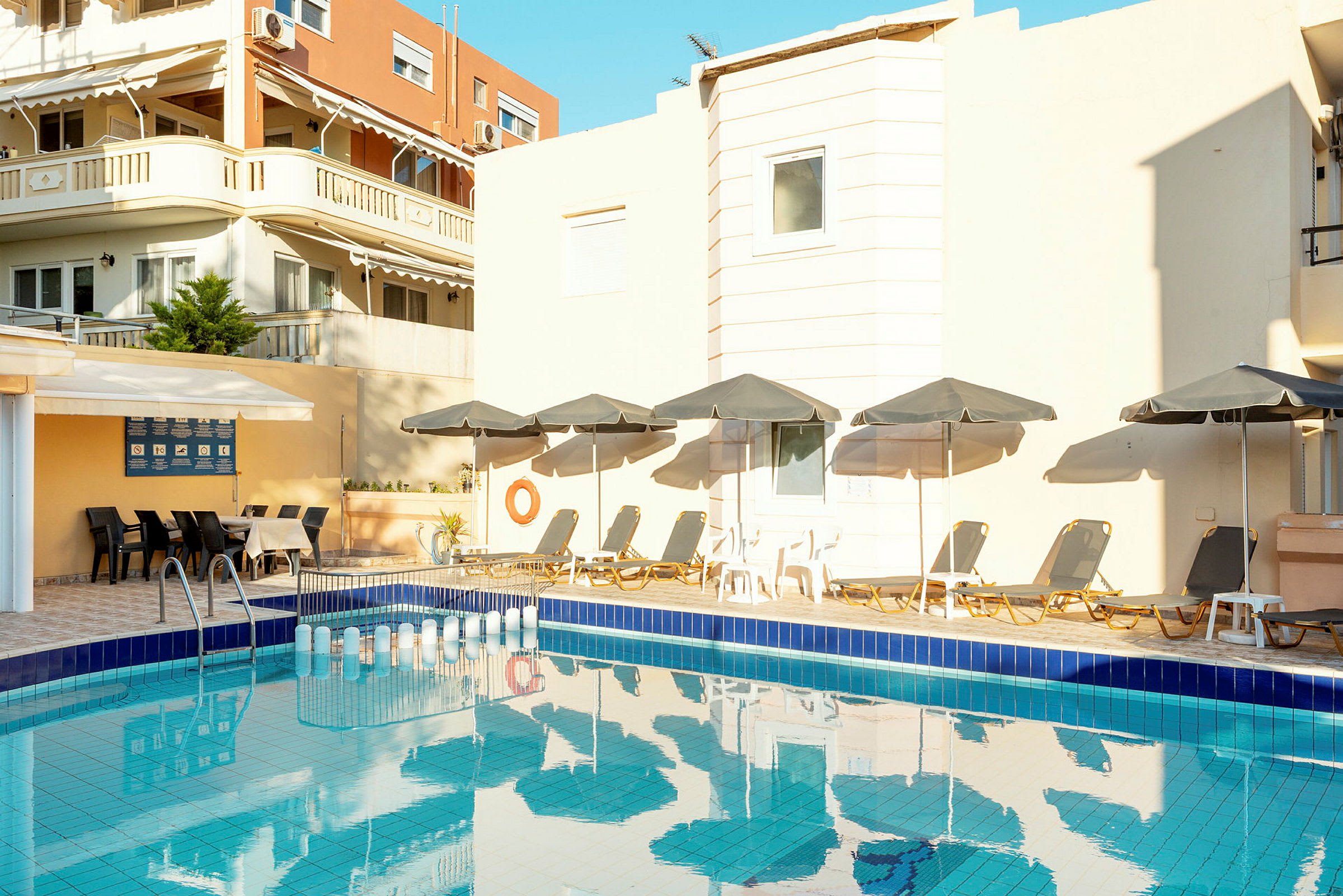 En swimmingpool omgiven av solstolar med parasoller och en byggnad i bakgrunden. (AI-genererad text)