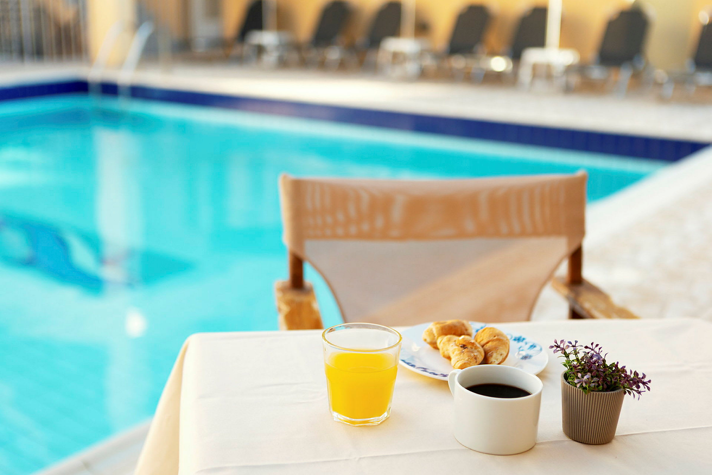 En bordduk med en frukostuppsättning vid en pool, innehållande juice, kaffe, croissanter och en liten växt. (AI-genererad text)