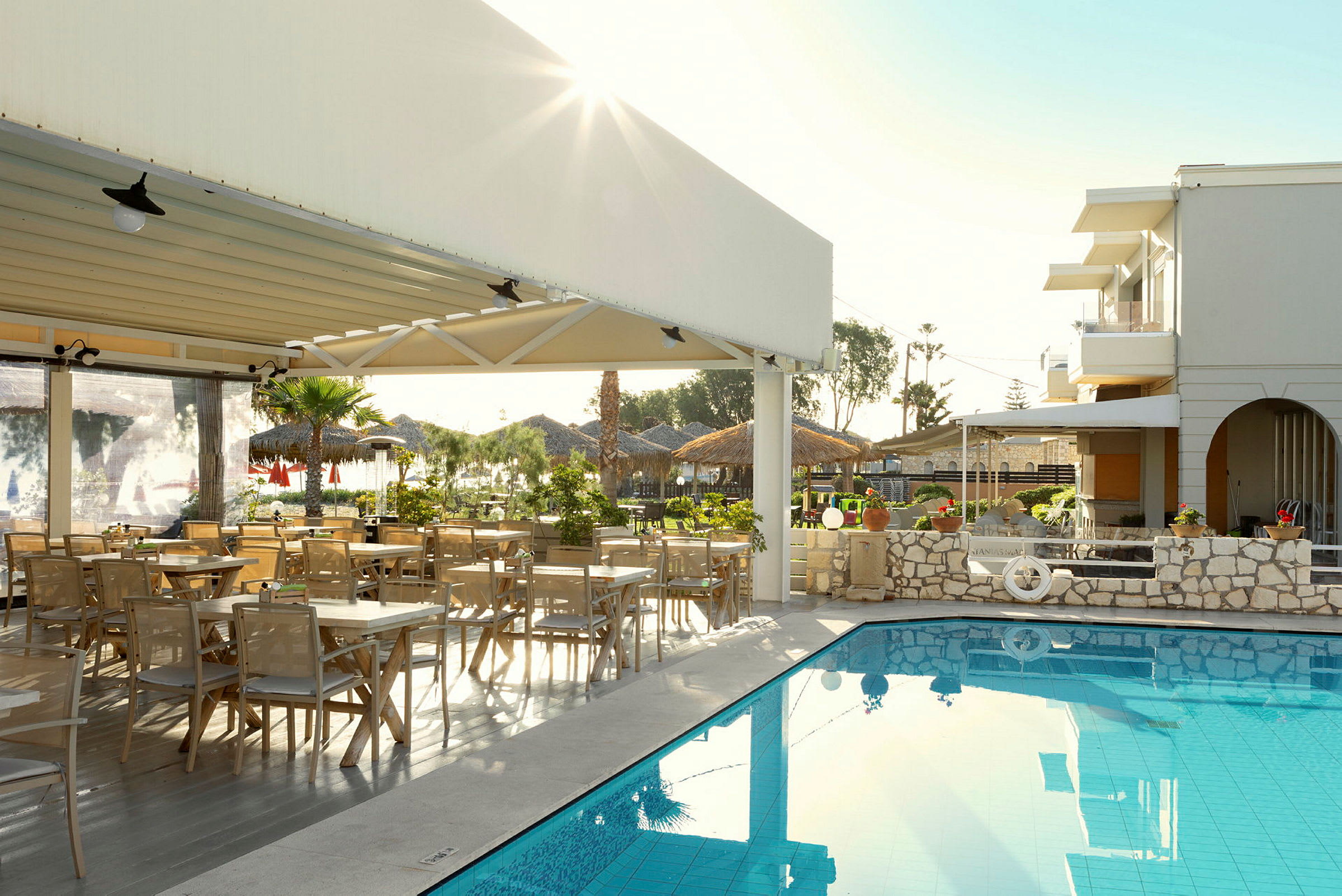 Utomhus miljö vid en restaurang med sittplatser och en pool. Byggnader och grönska syns i bakgrunden. (AI-genererad text)