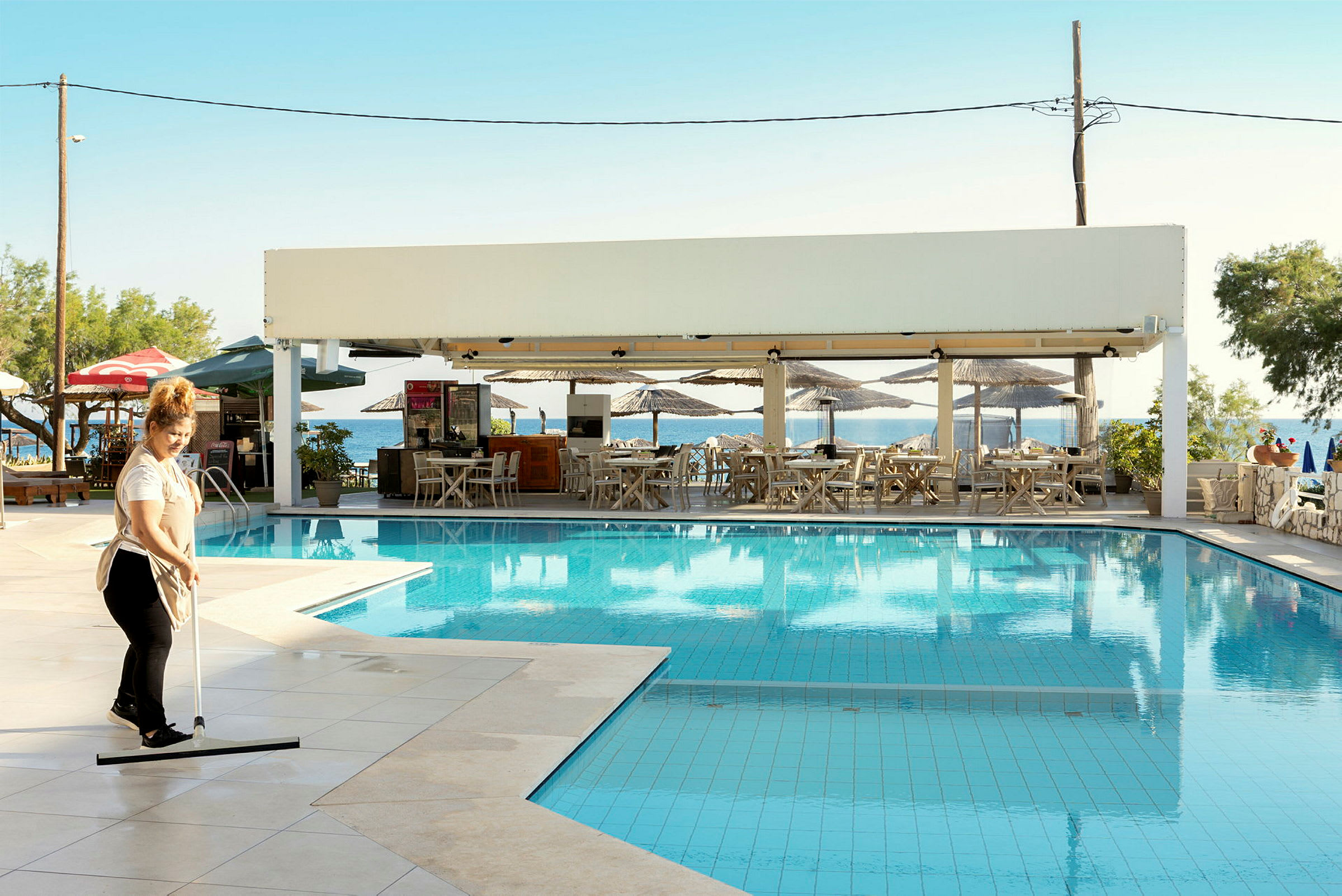 En kvinna rengör området vid en pool nära en utomhusrestaurang med solskydd. Restaurangen ligger vid havet. (AI-genererad text)