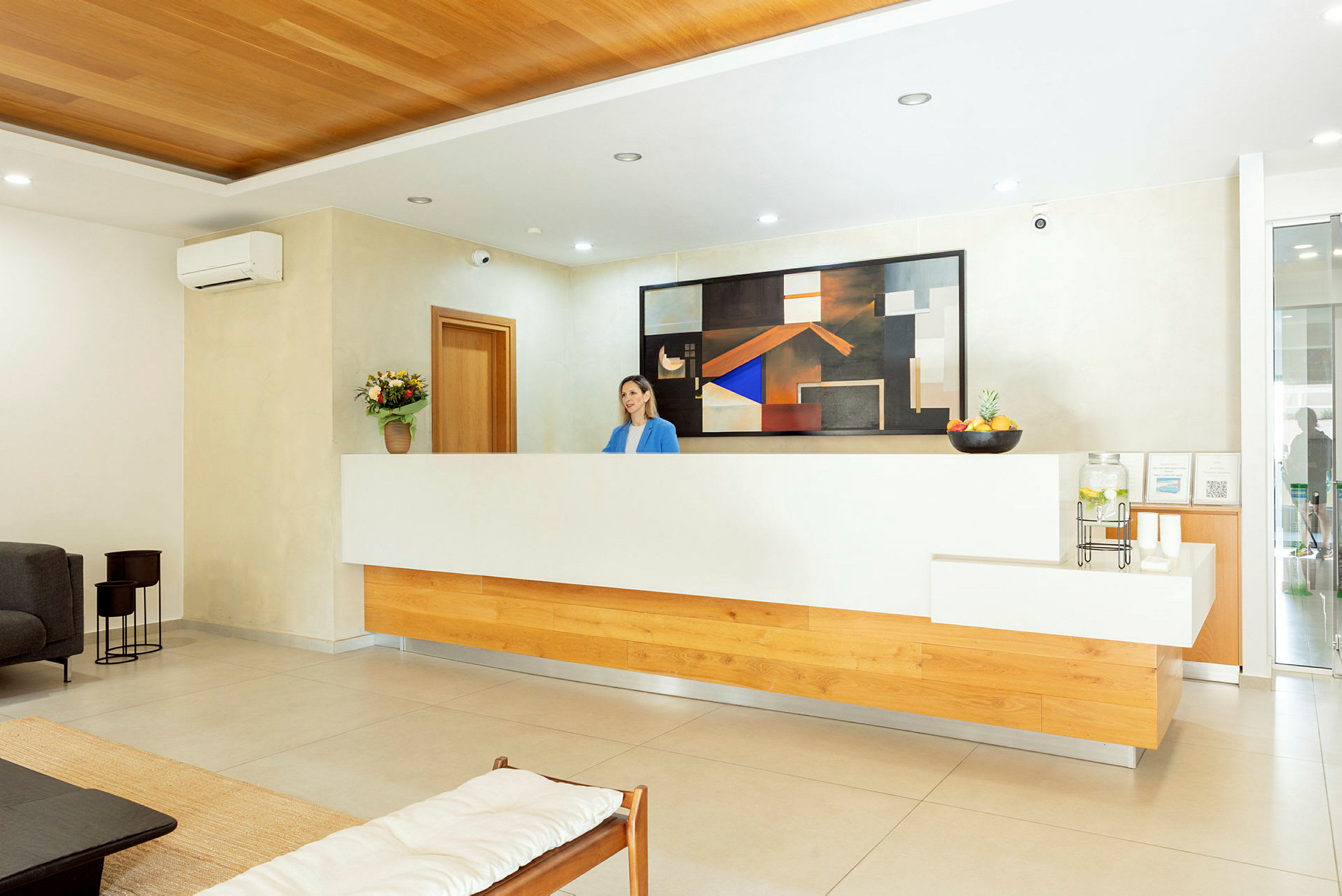 Ett hotellreceptionsområde med en receptionsdisk, en person bakom den, modern inredning och en tavla på väggen. (AI-genererad text)