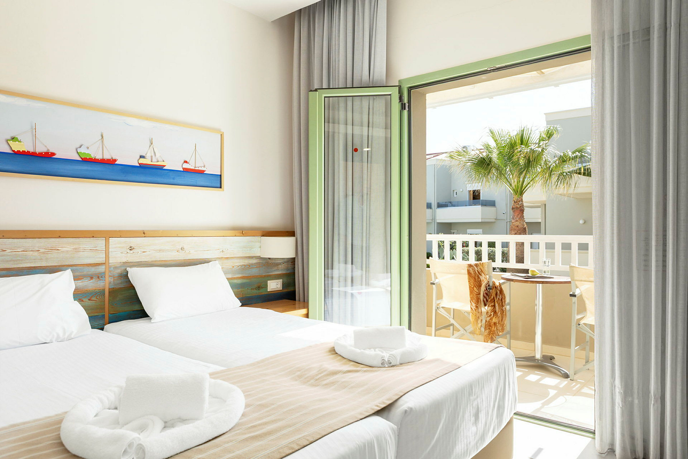 Ett ljust hotellrum med två enkelsängar, vita sängkläder och beige överkast. Ett inslag av båtmotiv hänger på väggen ovanför sängen. Rummet har en öppen balkongdörr med utsikt över en palm och ett bord med två stolar. (AI-genererad text)