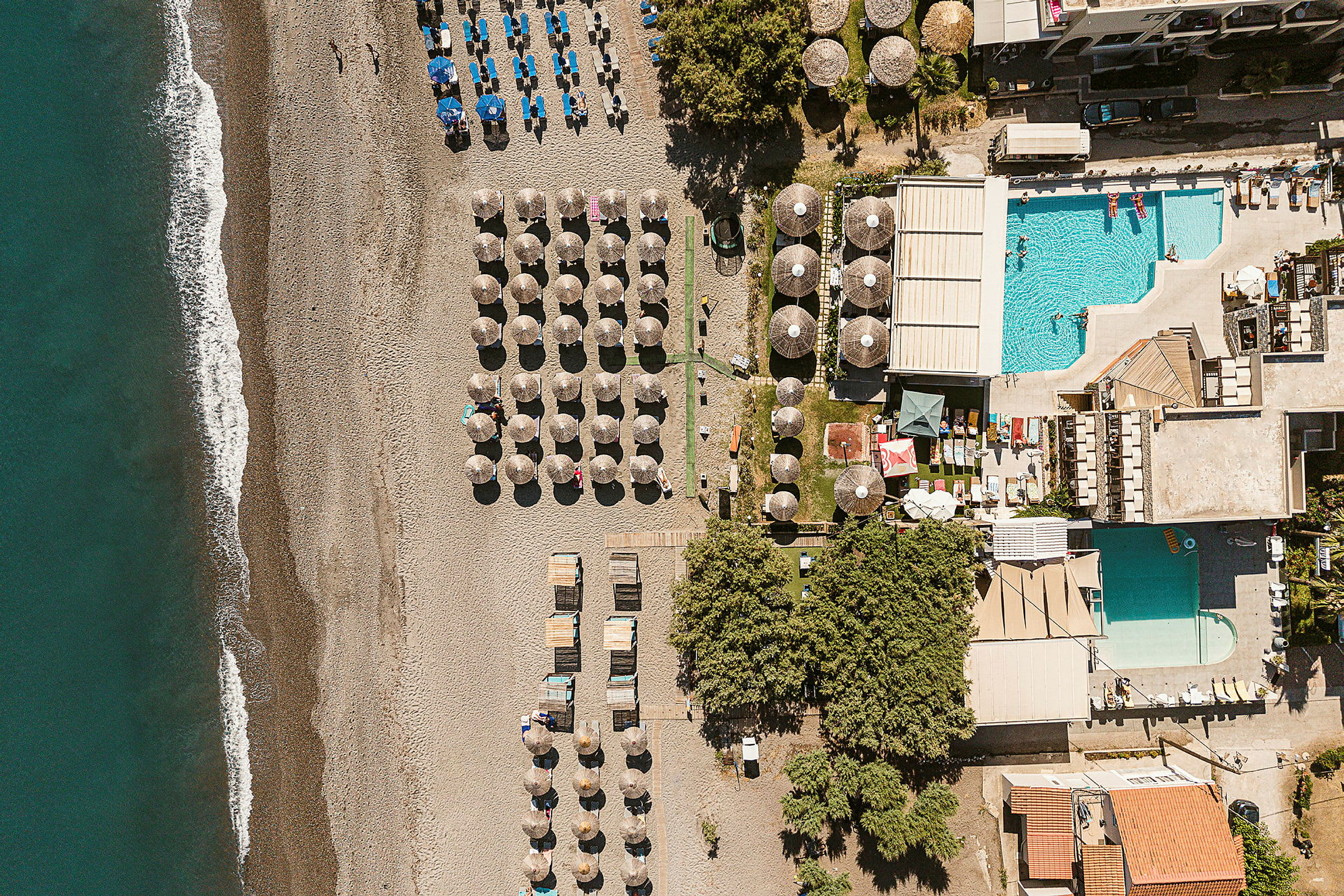 Flygfoto av en strand med solstolar och parasoller, bredvid en byggnad med en pool. (AI-genererad text)