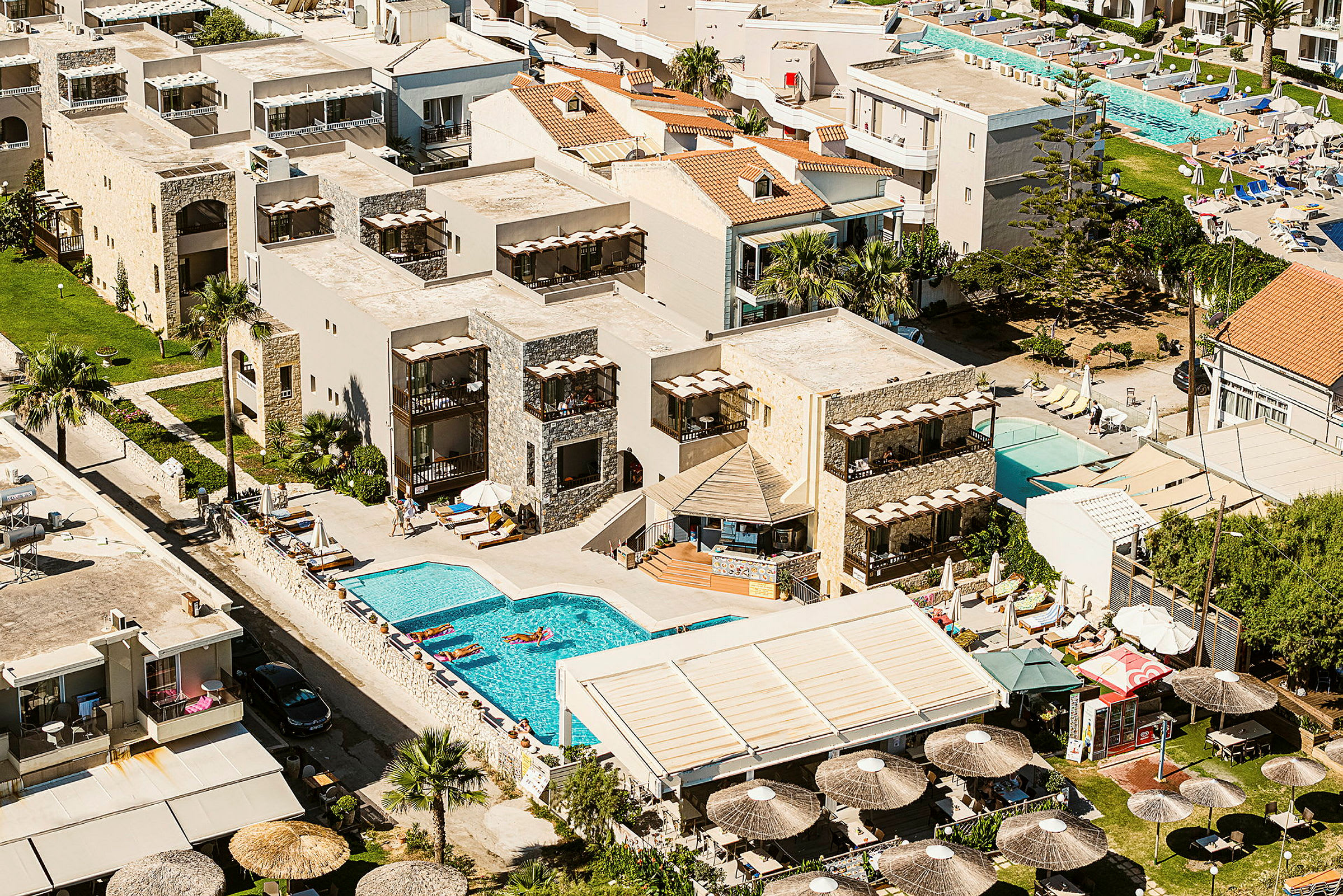 Flygfoto över ett hotellkomplex med pool och omgivande solstolar och byggnader. (AI-genererad text)