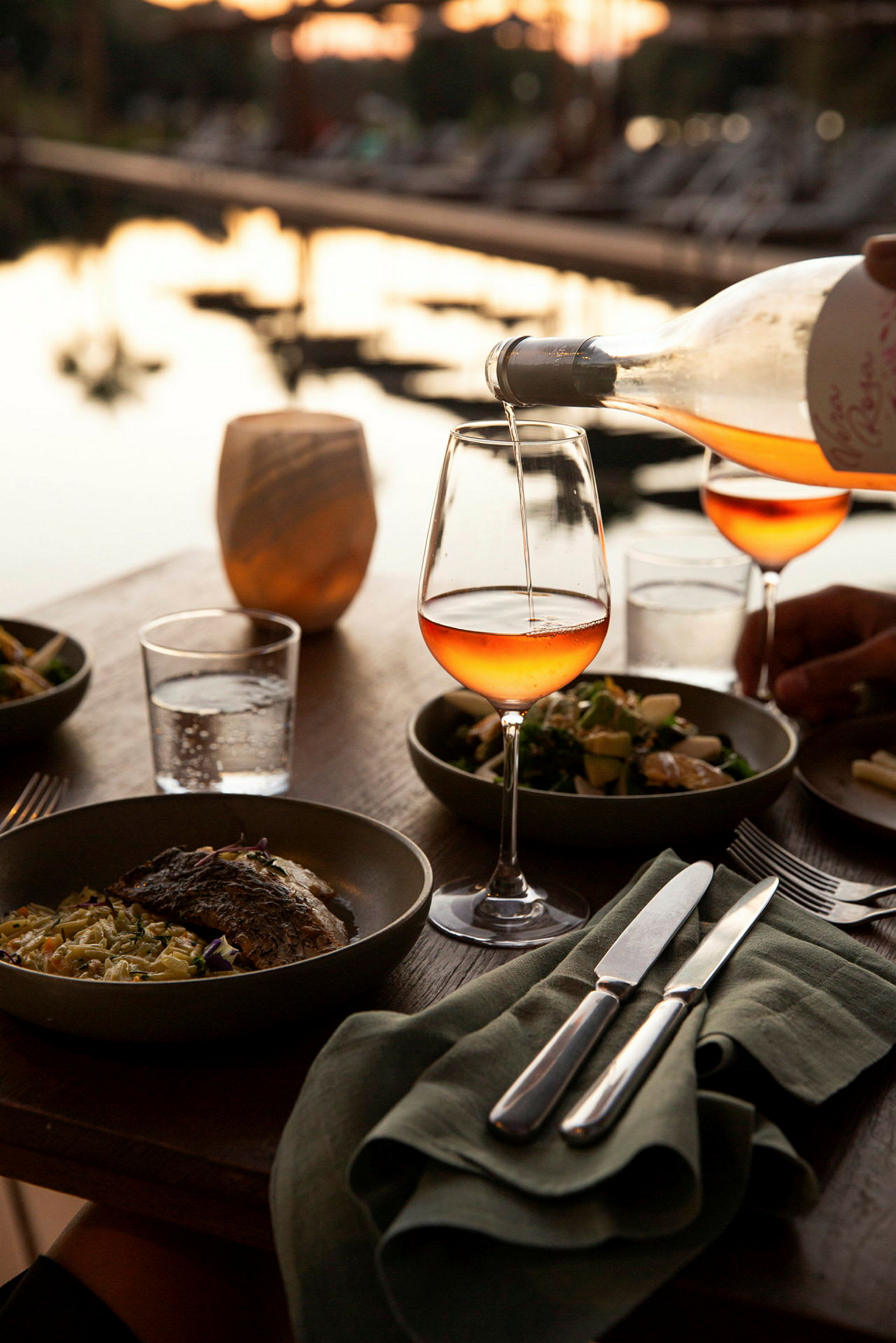 En vinflaska häller rosévin i ett vinglas på ett dukat bord vid vattnet. (AI-genererad text)