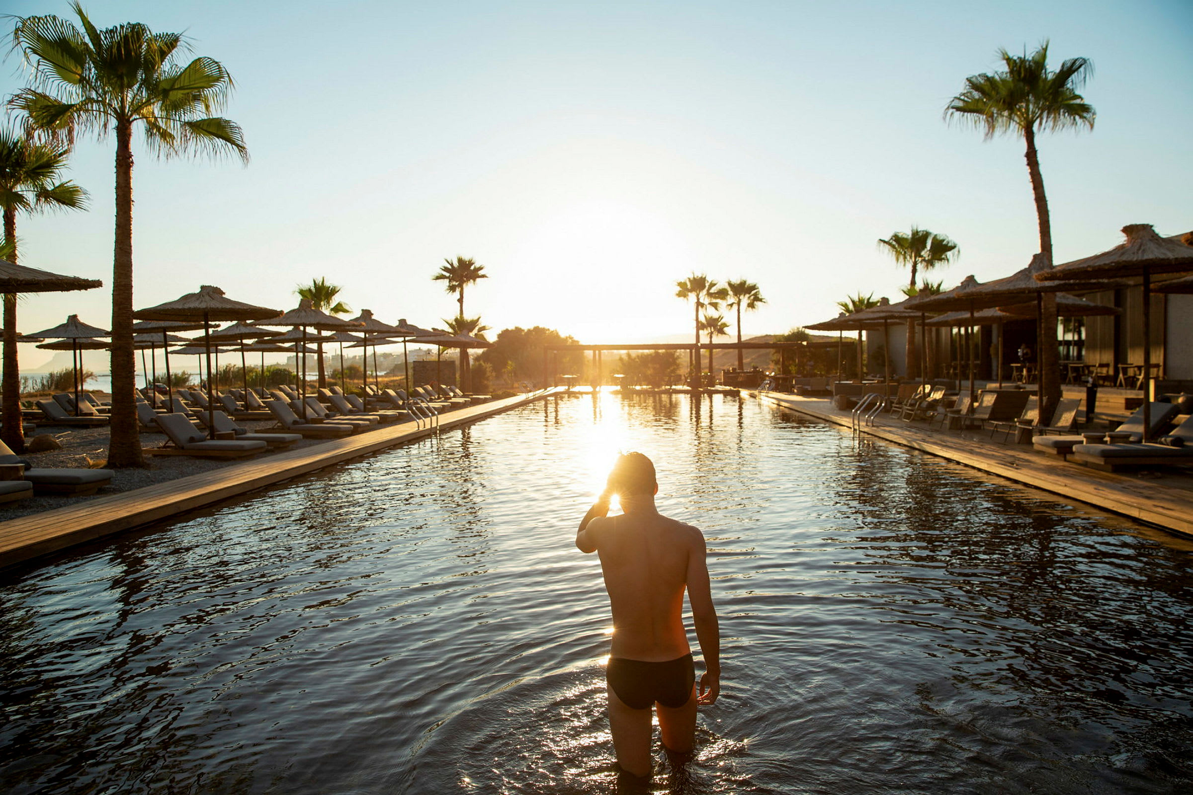 Person som går i en pool vid solnedgången med solstolar och parasoller runtom. (AI-genererad text)