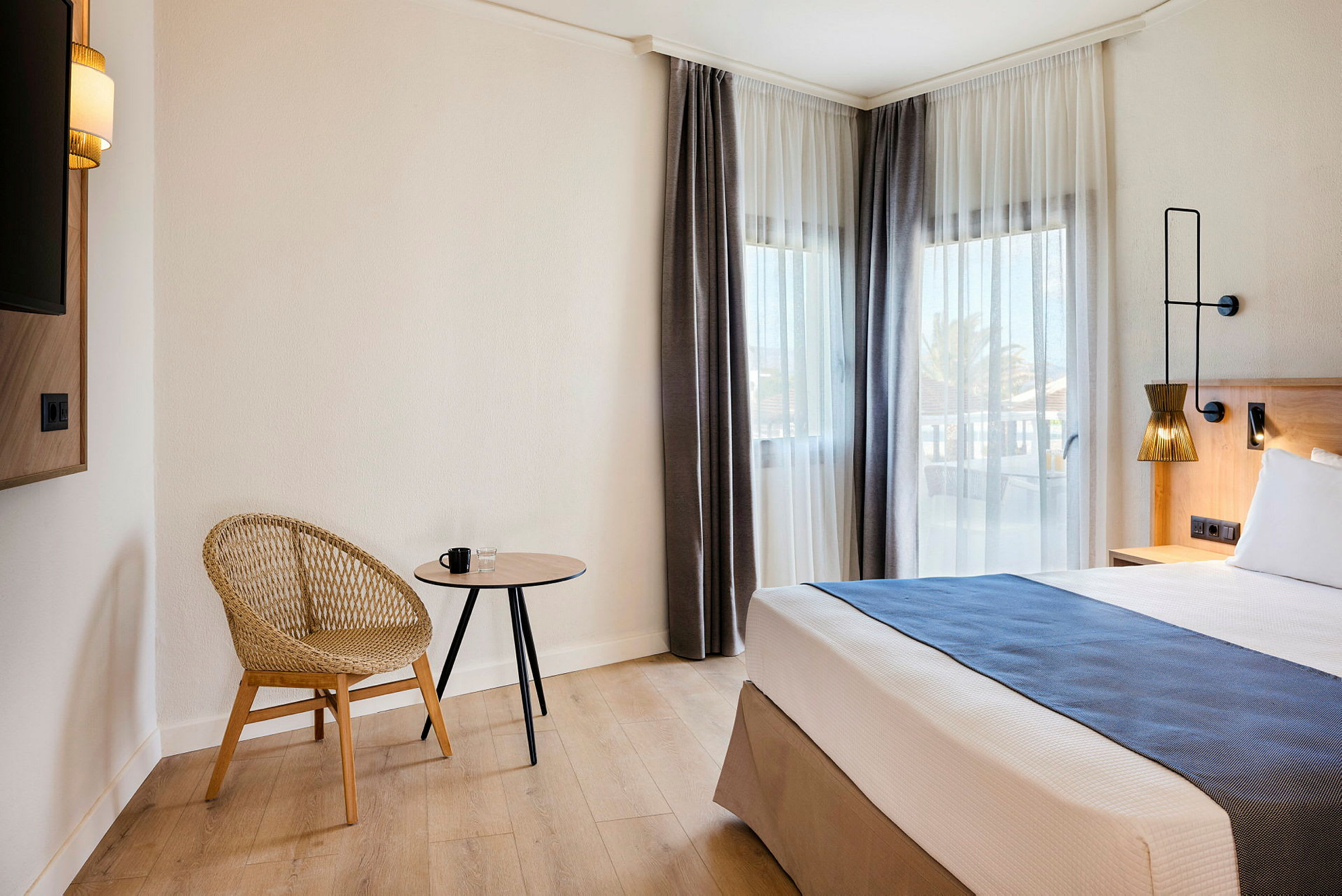 Ett hotellrum med en modern stol, ett litet bord och en säng bredvid fönster med ljusa gardiner. (AI-genererad text)