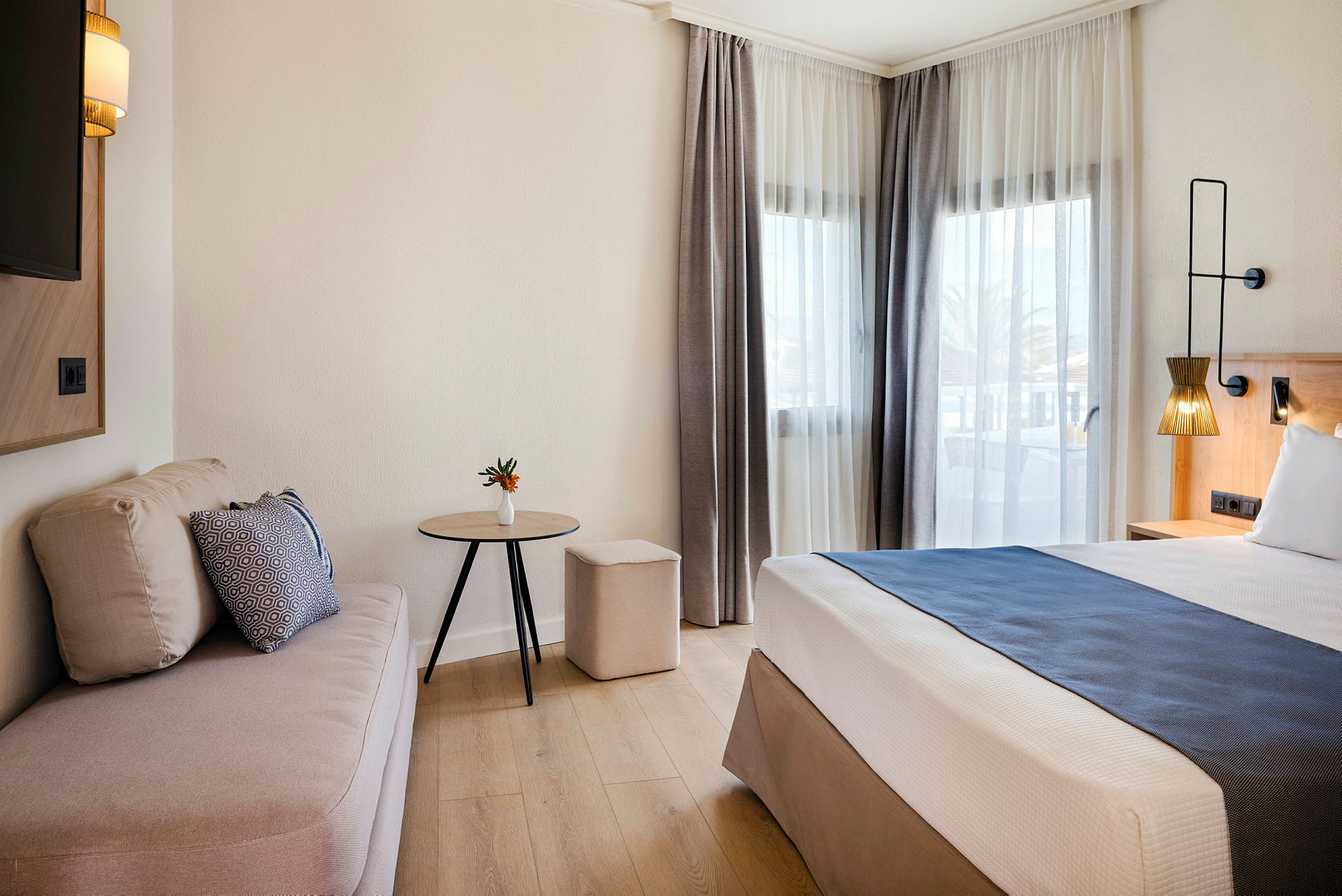 En hotellrum med en säng, soffa, och bord bredvid stora fönster. (AI-genererad text)