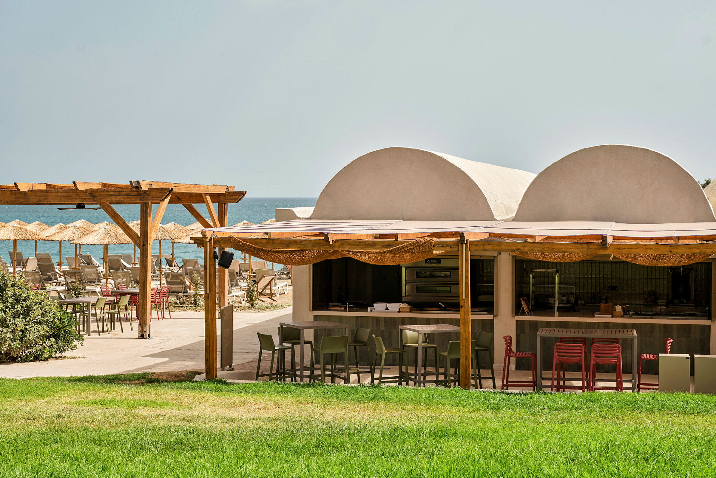 Utomhusrestaurang vid stranden med grön gräsmatta, träpergola och parasoller. (AI-genererad text)