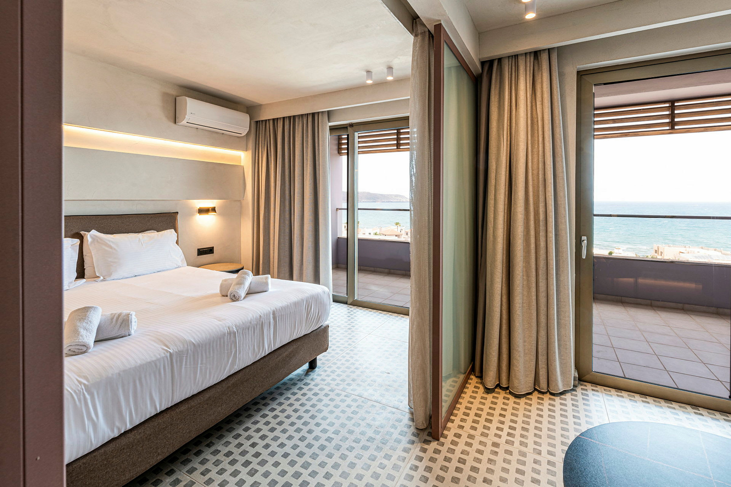 Hotellrum med säng och handdukar, stora glasdörrar som leder ut till en balkong som visar havet. (AI-genererad text)