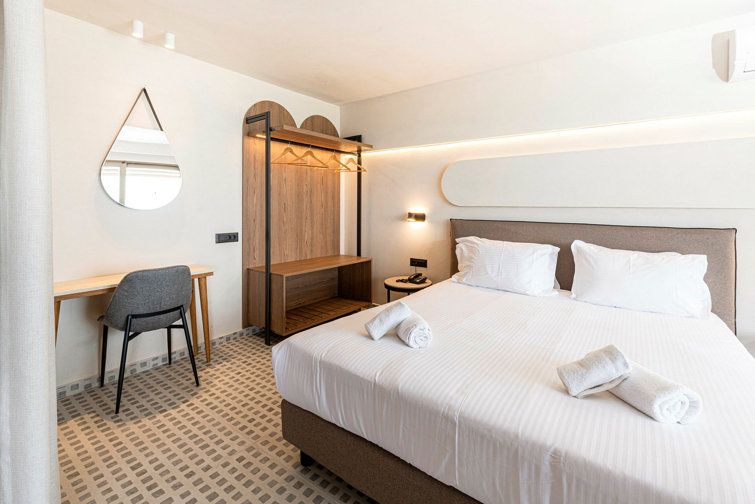 Ett hotellrum med en uppbäddad säng, skrivbord med stol, väggspegel och öppet klädskåp. (AI-genererad text)