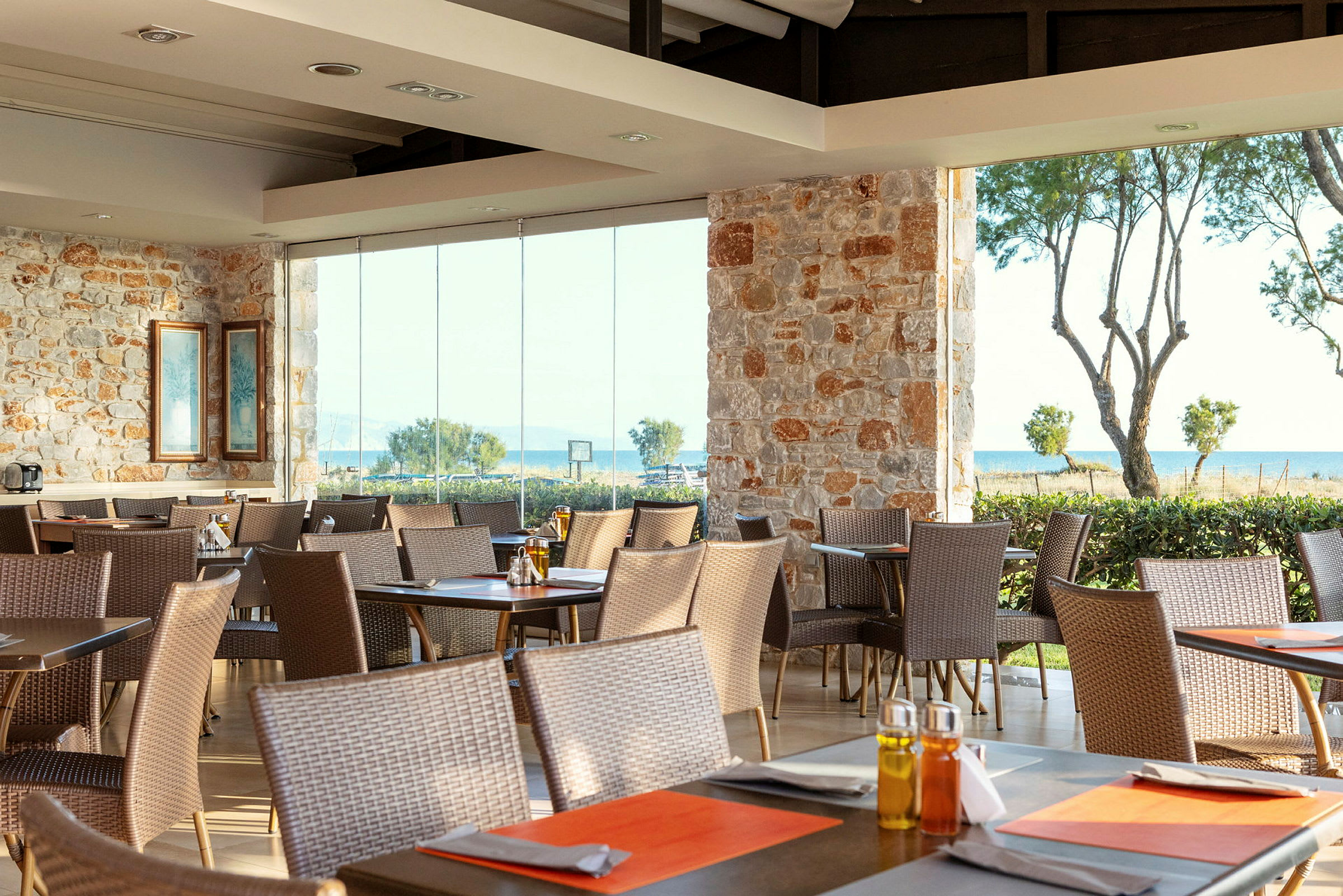 Restaurang med stenväggar och stora fönster som vetter mot havet, med flera bord och stolar. (AI-genererad text)