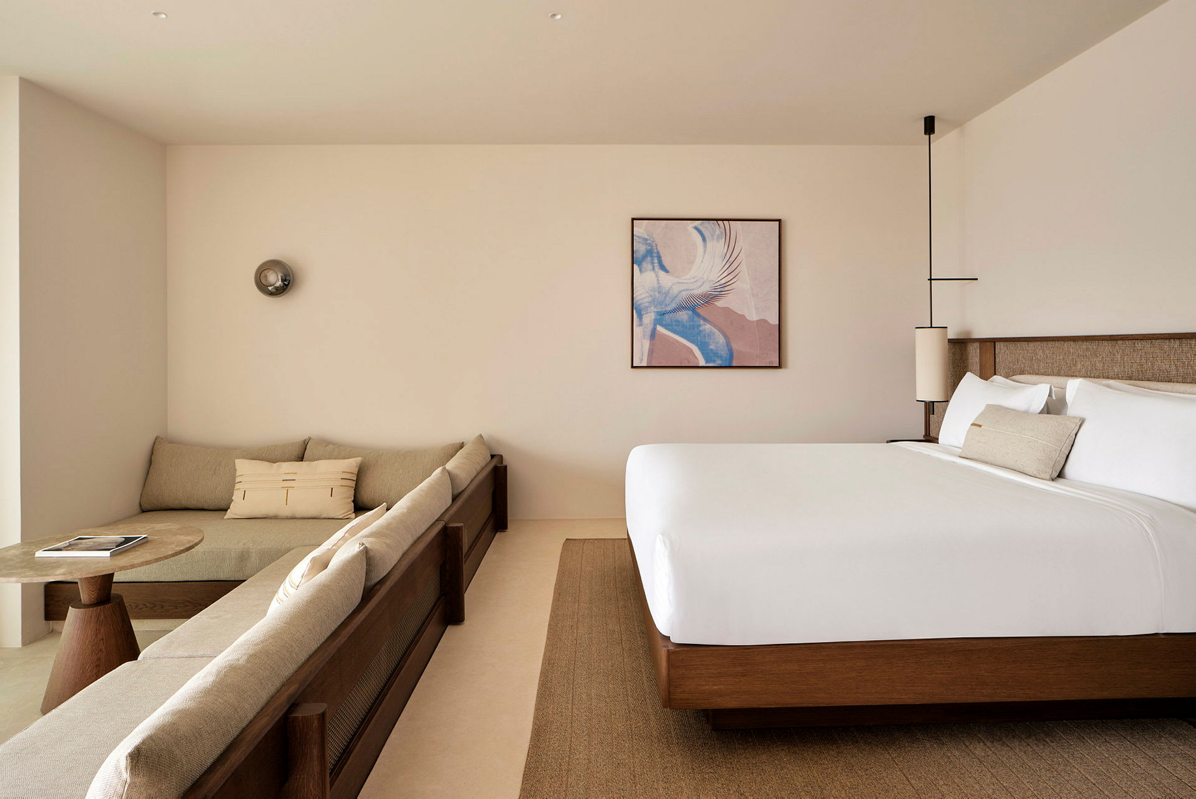 Ett modernt hotellrum med beige soffa, ett runt soffbord och en stor säng. En tavla samt dekor lampor syns. (AI-genererad text)