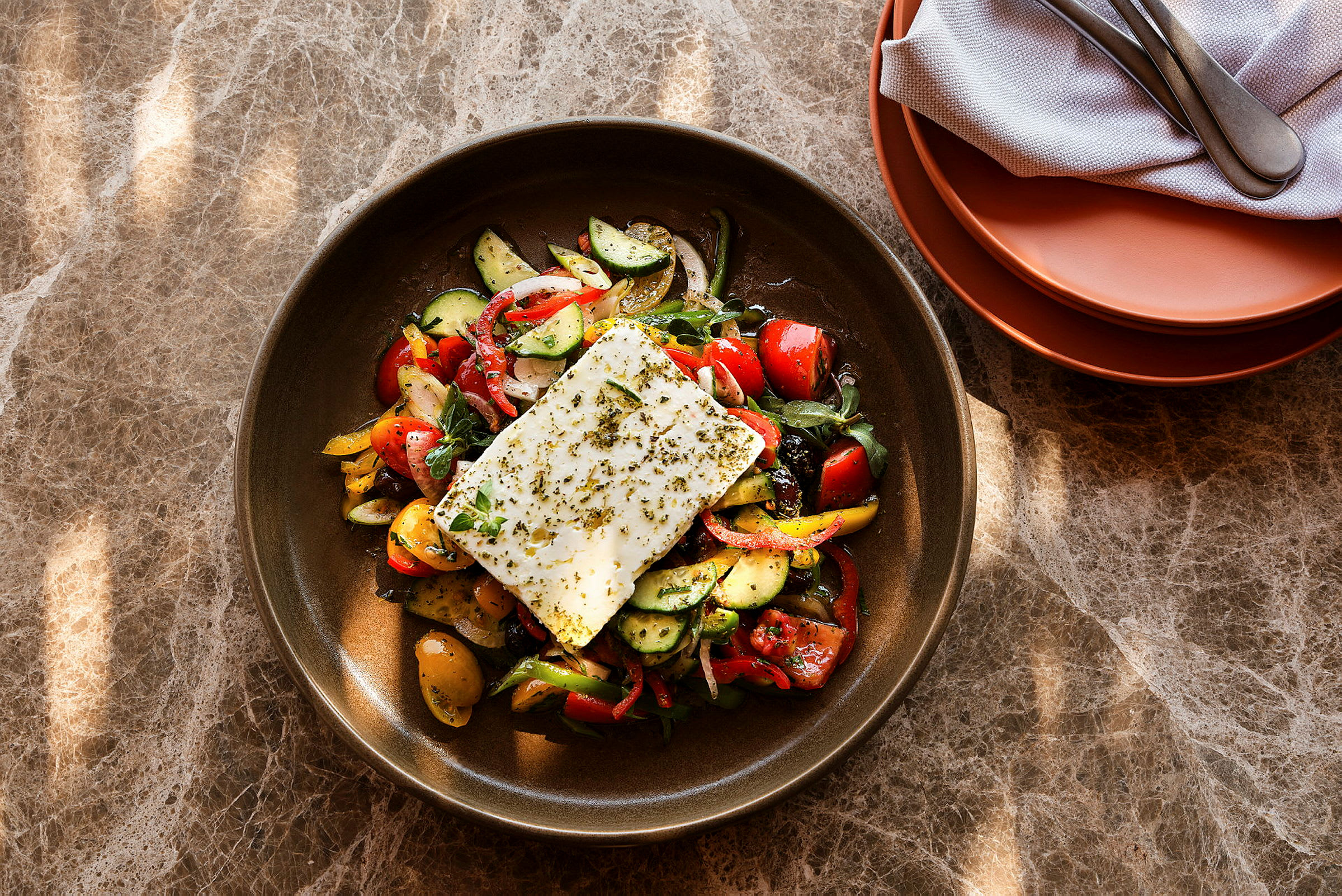 En tallrik med en sallad av grönsaker och en stor bit vitost ovanpå. Tallriken står på ett marmorbord. (AI-genererad text)