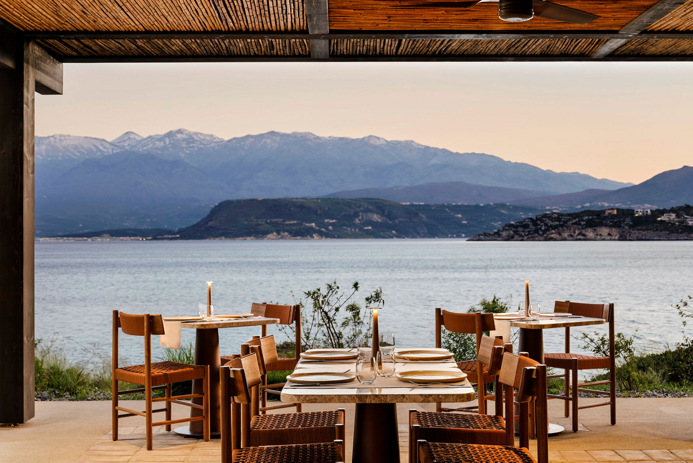 En utomhusrestaurang med dukade bord, med utsikt över havet och berg i bakgrunden. (AI-genererad text)