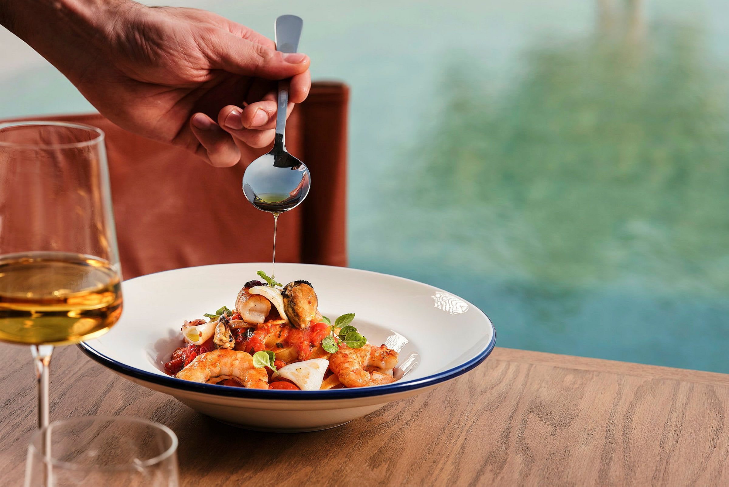 En hand som håller en sked med olja droppar över en maträtt med skaldjur och tomatsås, bredvid ett glas vin, ståendes på ett bord nära vatten. (AI-genererad text)