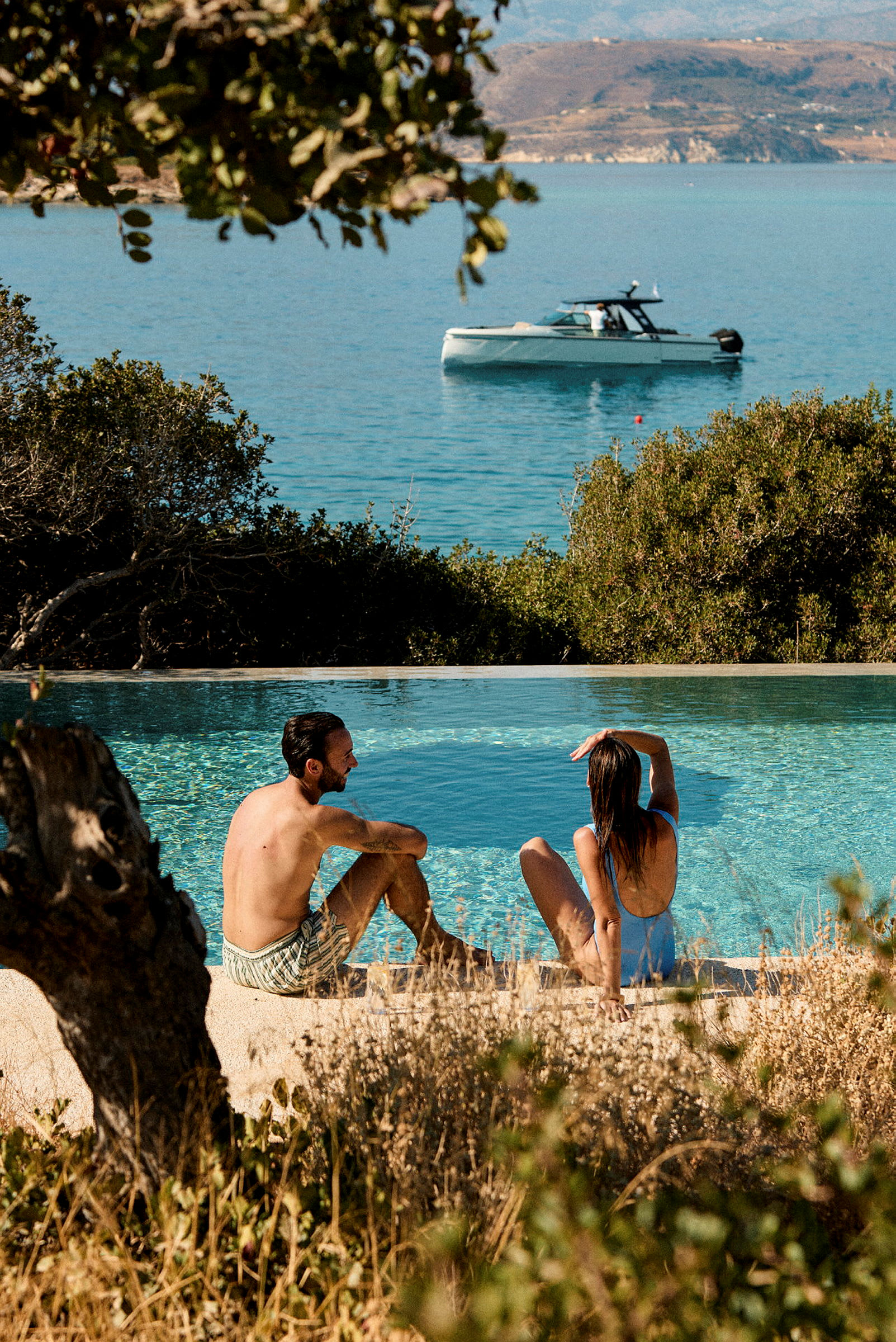 Två personer som sitter vid kanten av en pool med utsikt över havet där en båt flyter. (AI-genererad text)