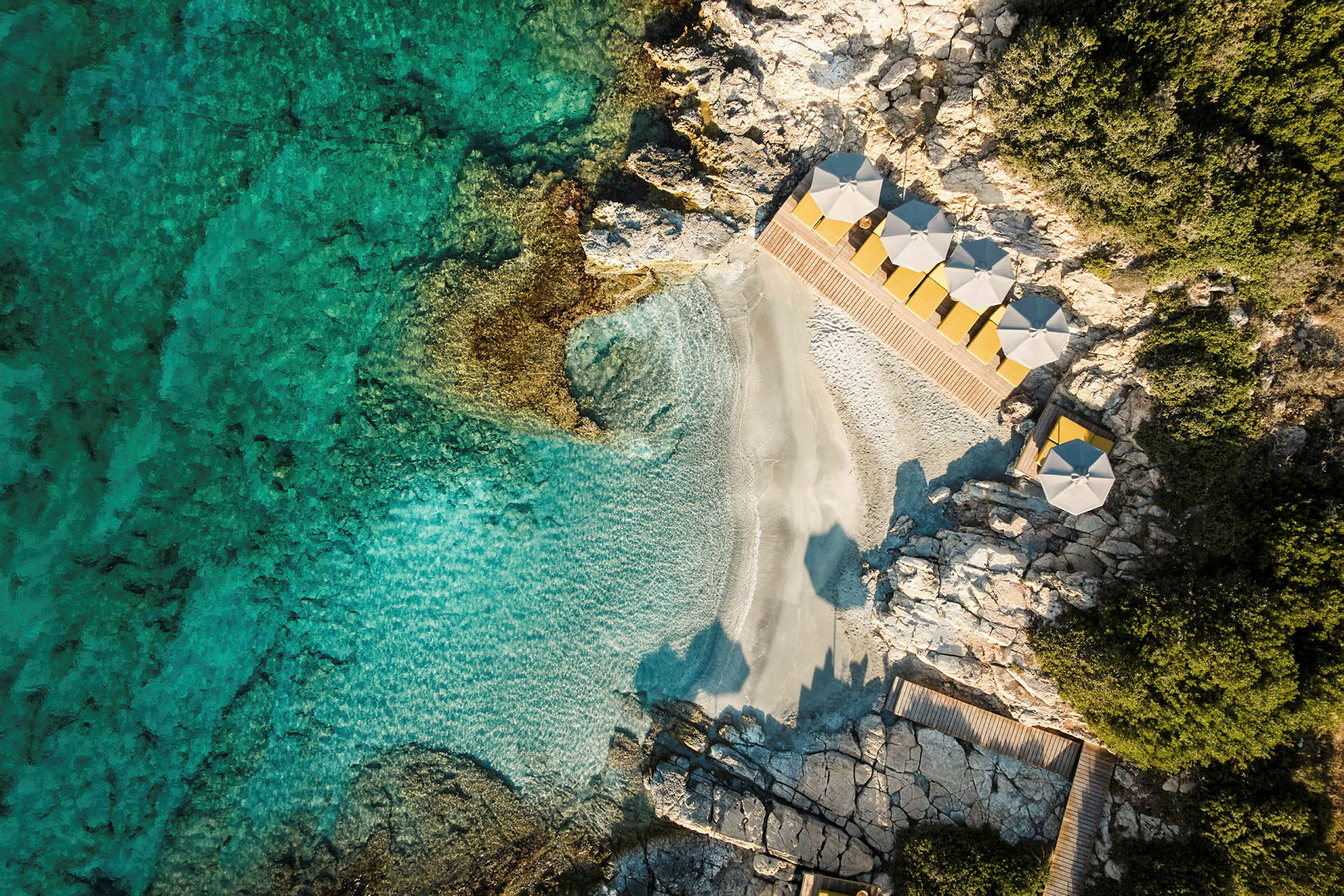 Flygbild över en liten strand med klart turkost vatten, omgiven av klippor och med parasoller på strandkanten. (AI-genererad text)