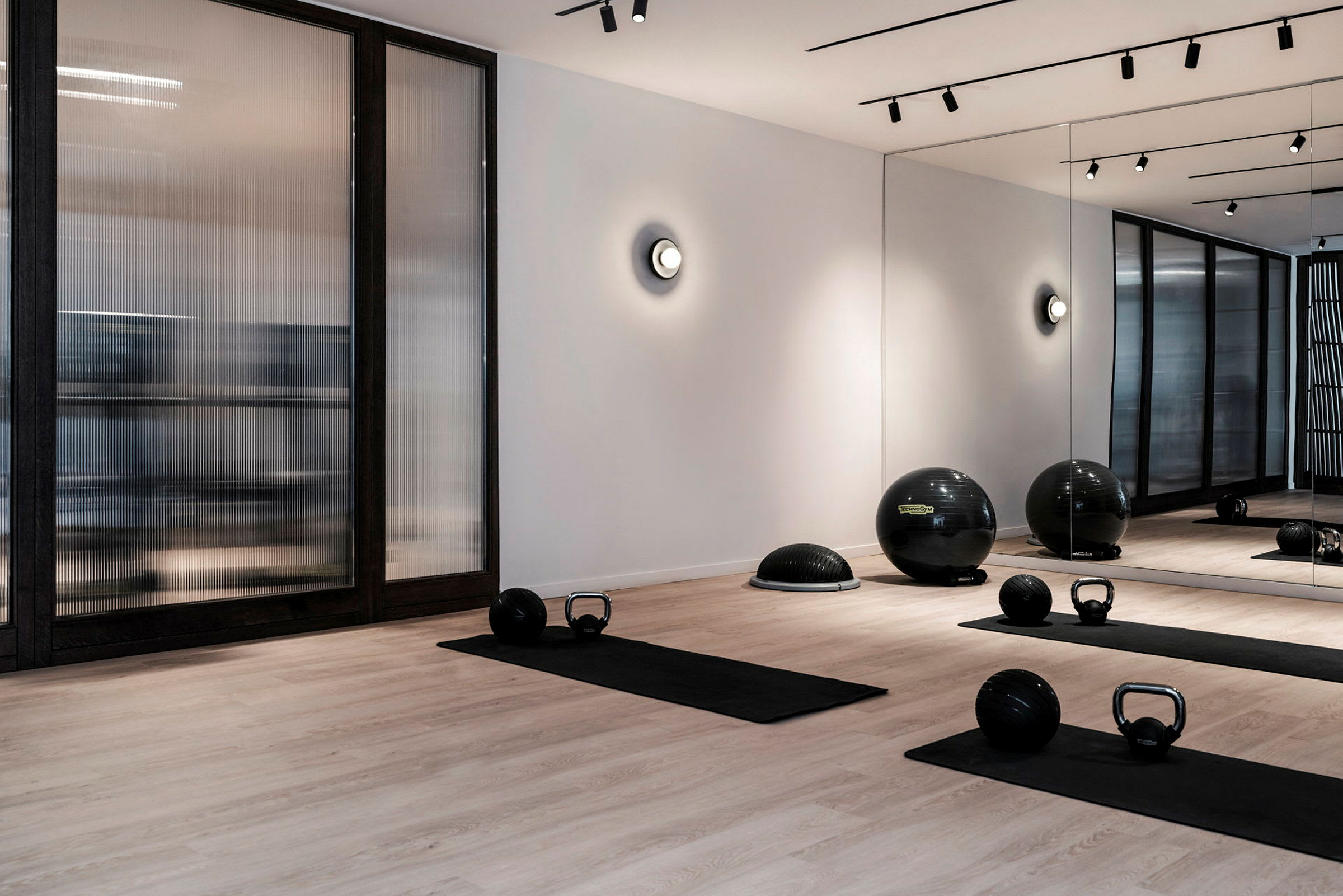 Ett modernt gym med träningsmattor, kettlebells och fitnessbollar på ett trägolv. Väggen har speglar och glaspartier. (AI-genererad text)