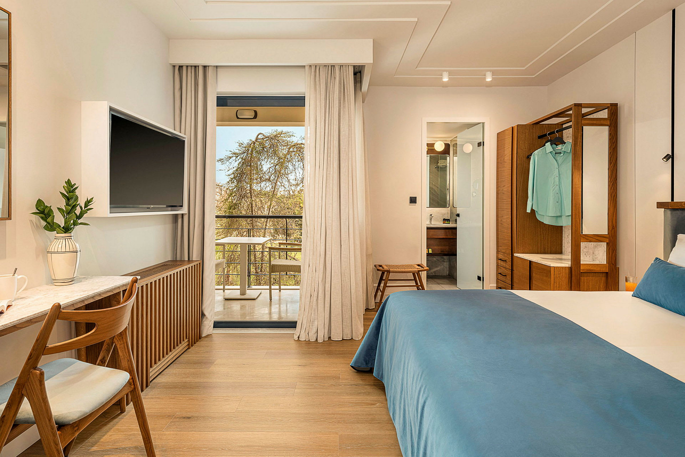 Hotellrum med dubbelsäng, skrivbord och TV. Glasdörrar leder ut till en balkong. Badrum synligt genom en öppen dörr. (AI-genererad text)