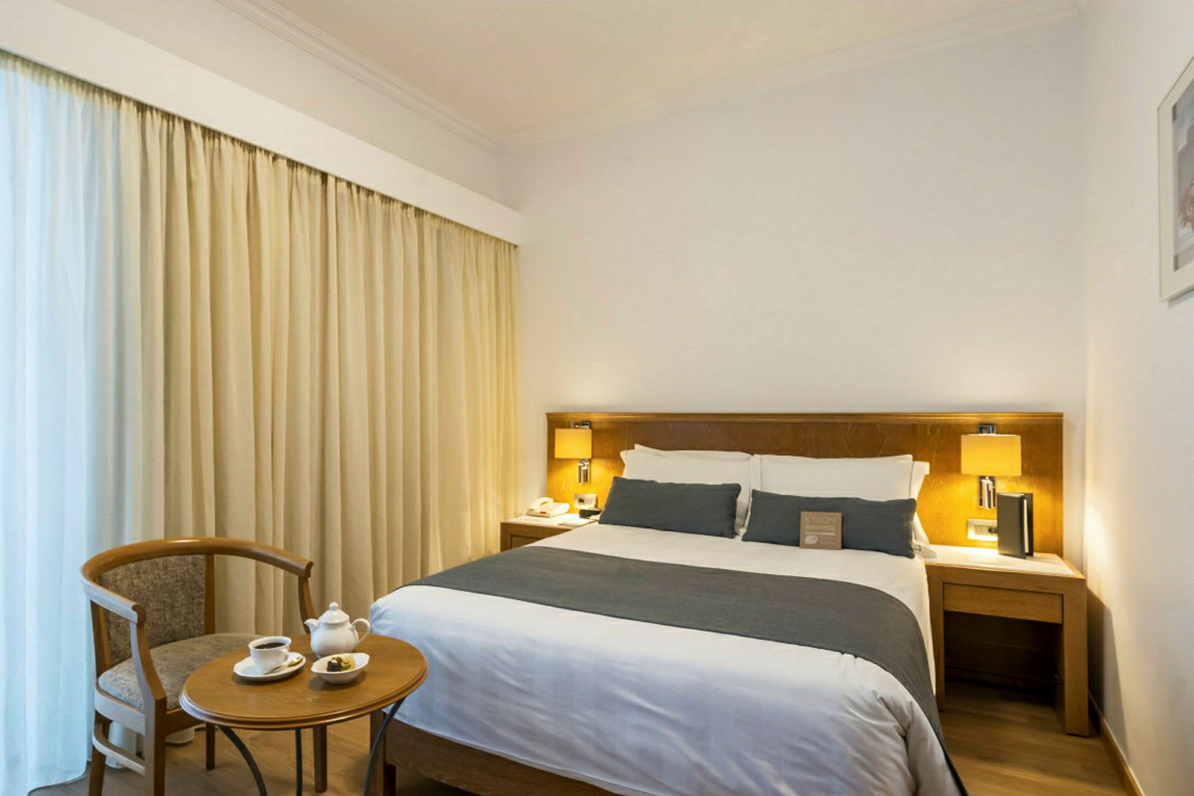 Ett hotellrum med en dubbelsäng, sängbord och lampa samt ett litet bord med två koppar och en stol. (AI-genererad text)