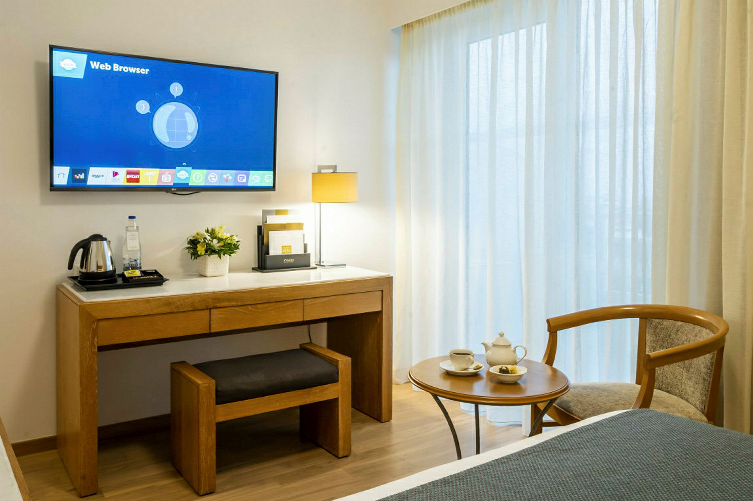 Ett hotellrum med en väggmonterad TV ovanför ett skrivbord med en stol och en vattenkokare på toppen. Bredvid skrivbordet finns ett sidobord med tekoppar och en stol. Bakgrunden innehåller fönster med ljusgenomsläppliga gardiner. (AI-genererad text)