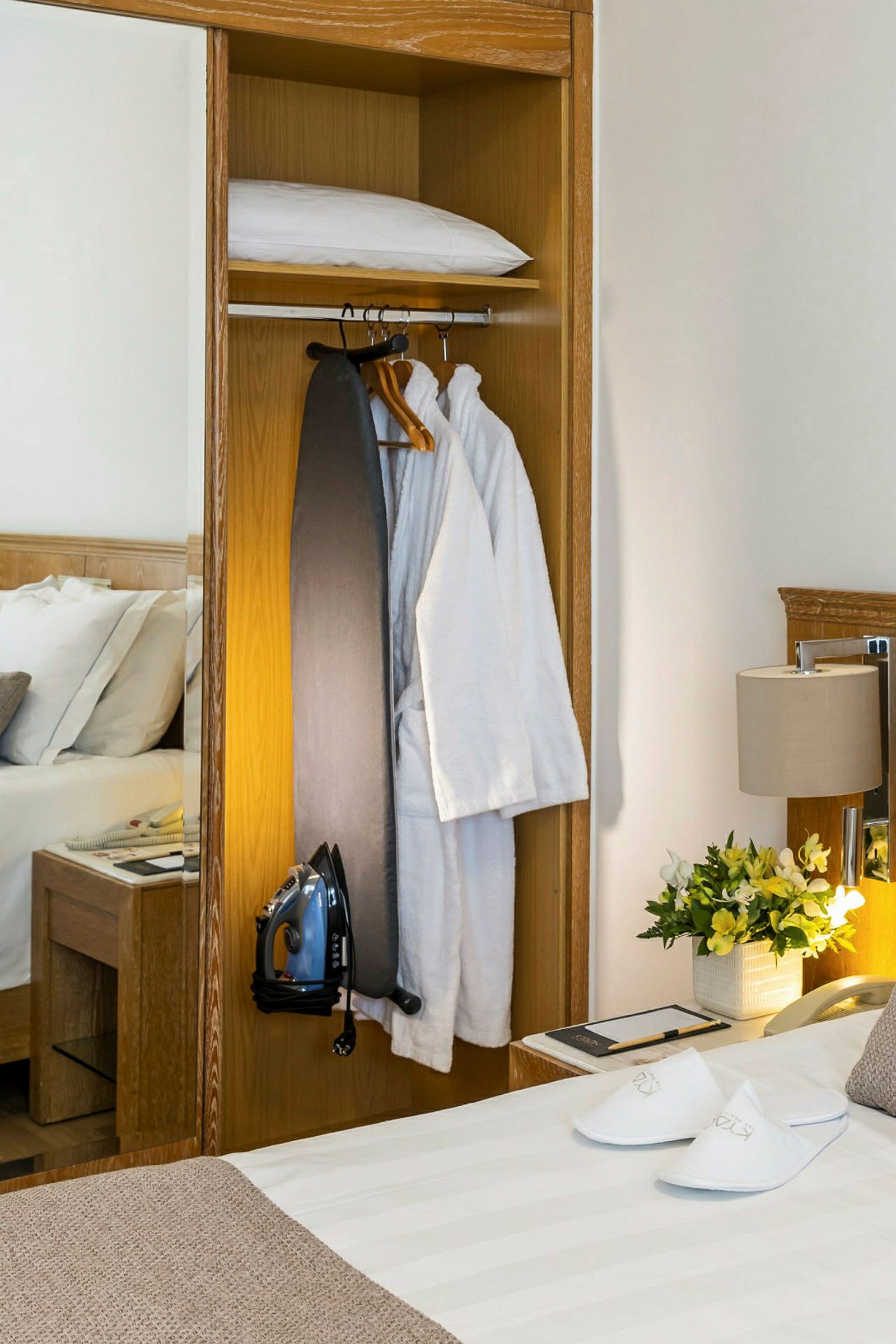 En garderob med klädhängare, en strykbräda, en strykjärn och badrockar. Till vänster syns en säng och till höger lampor och blommor. (AI-genererad text)