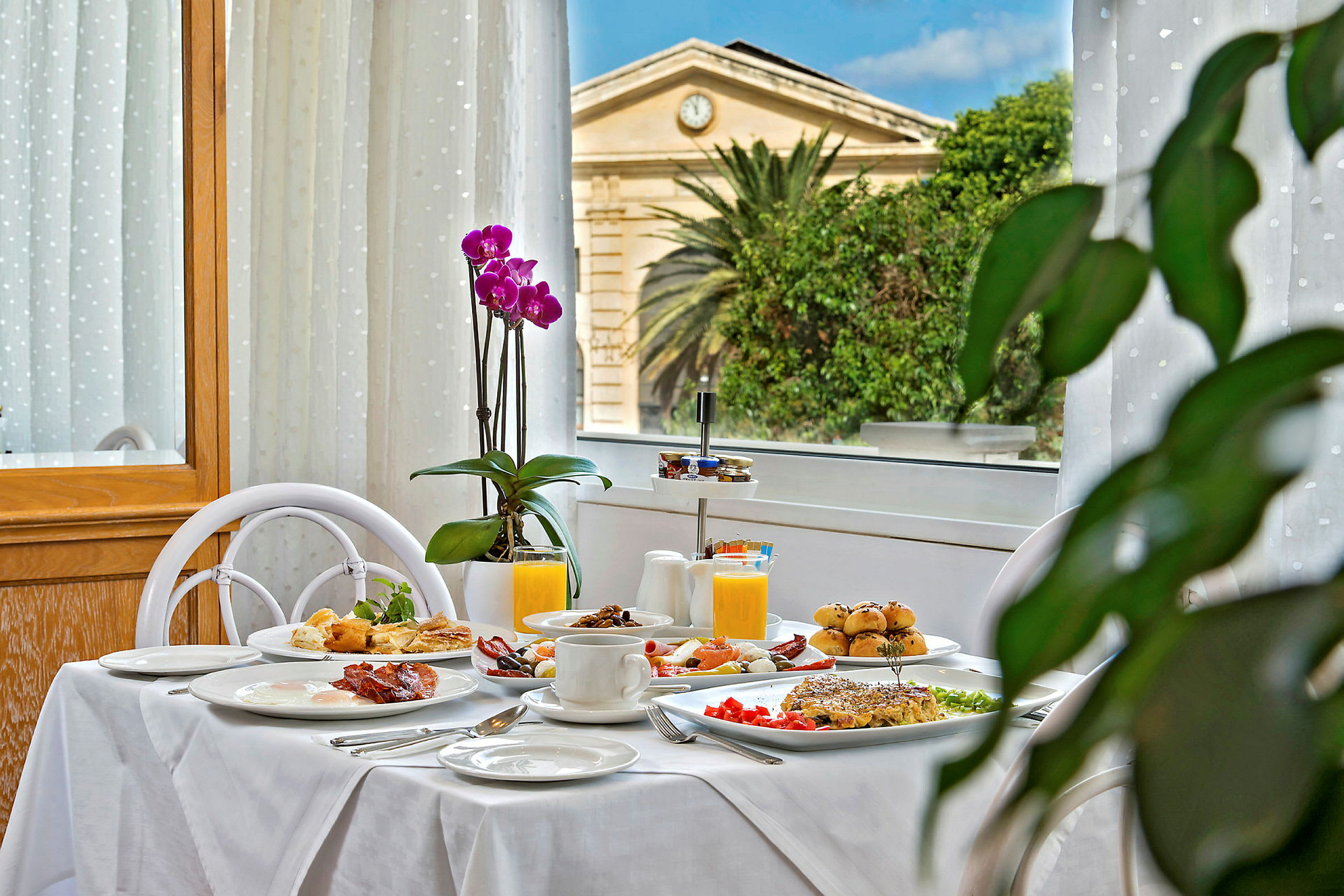 Ett dukat frukostbord med olika rätter som juice, kaffe och bakverk. I bakgrunden syns en byggnad, växtlighet och blå himmel. (AI-genererad text)