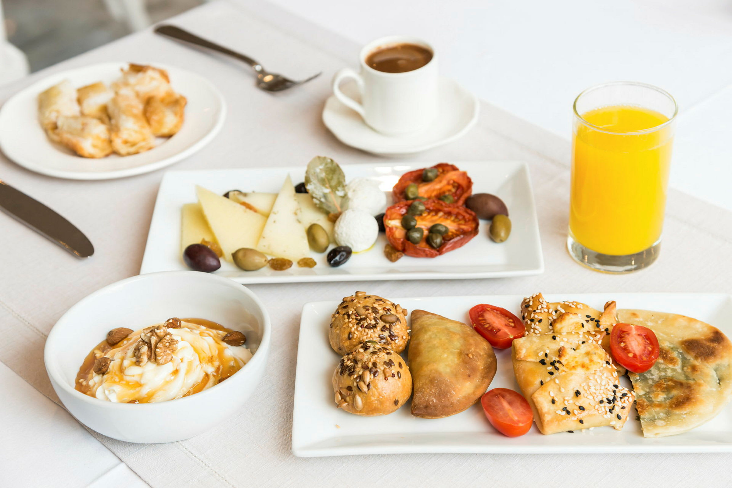 På bilden syns en frukostbeställning med olika ingredienser såsom bröd, ost, torkade tomater, oliver och kapris. Det finns dessutom en skål med yoghurt, några bakverk och ett glas apelsinjuice. (AI-genererad text)