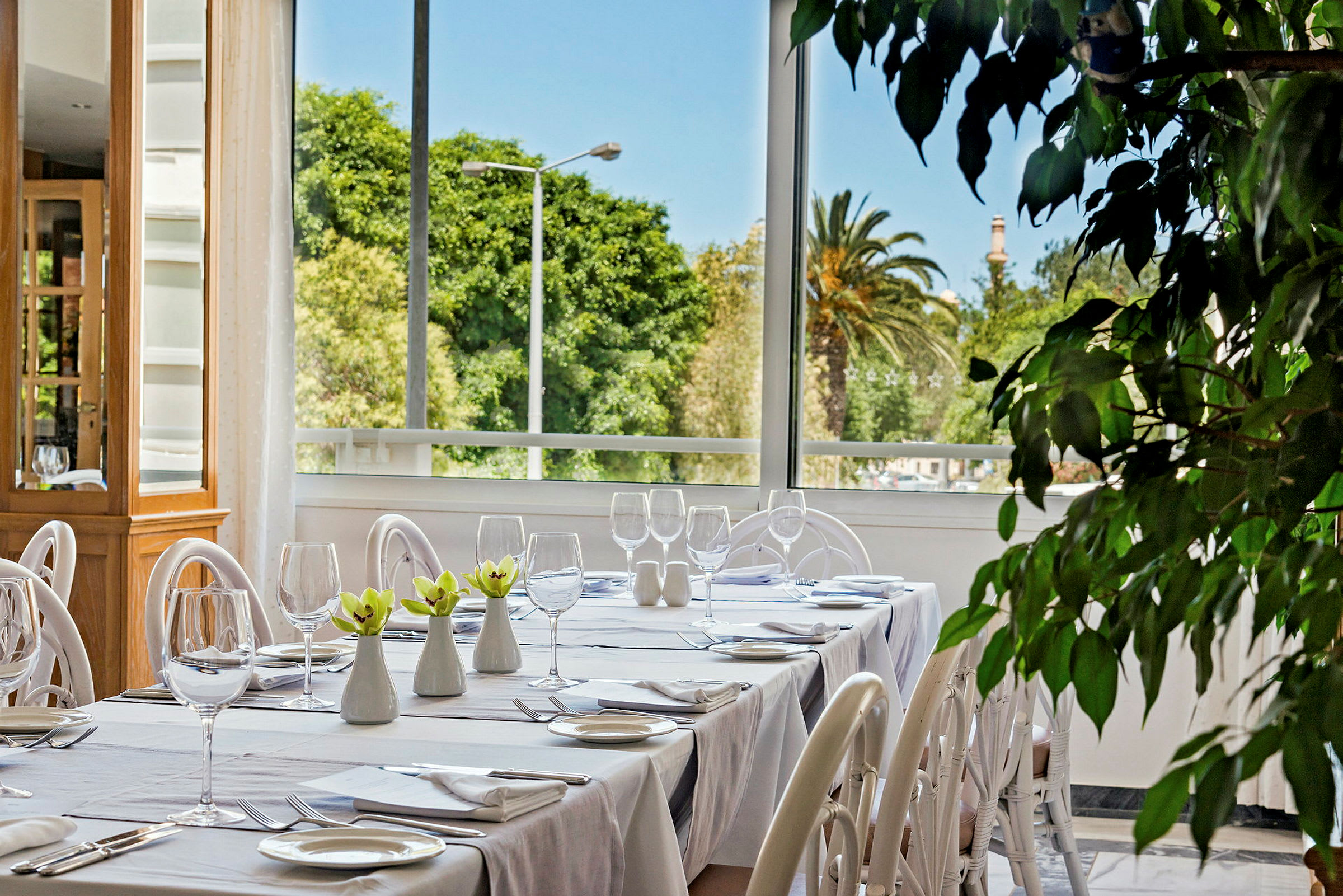 På bilden syns ett dukat bord i en restaurang med vit porslin och glas. I bakgrunden finns stora fönster som visar grönskande utsikt. (AI-genererad text)