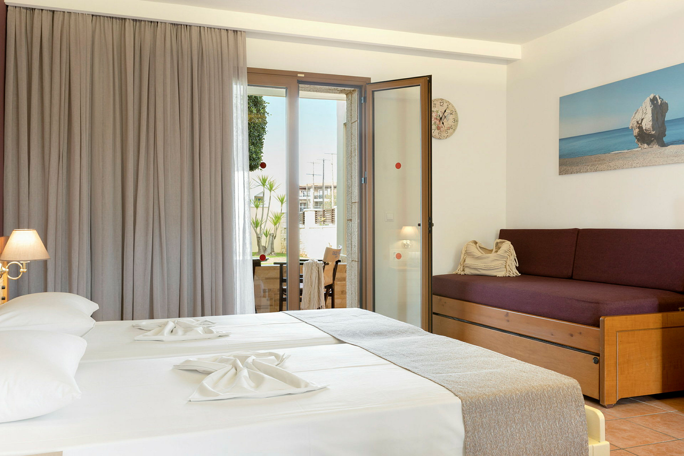 Ett hotellrum med två sängar klädda i ljusa lakan, en lila soffa med kuddar, och en glasdörr som leder ut till en balkong. (AI-genererad text)