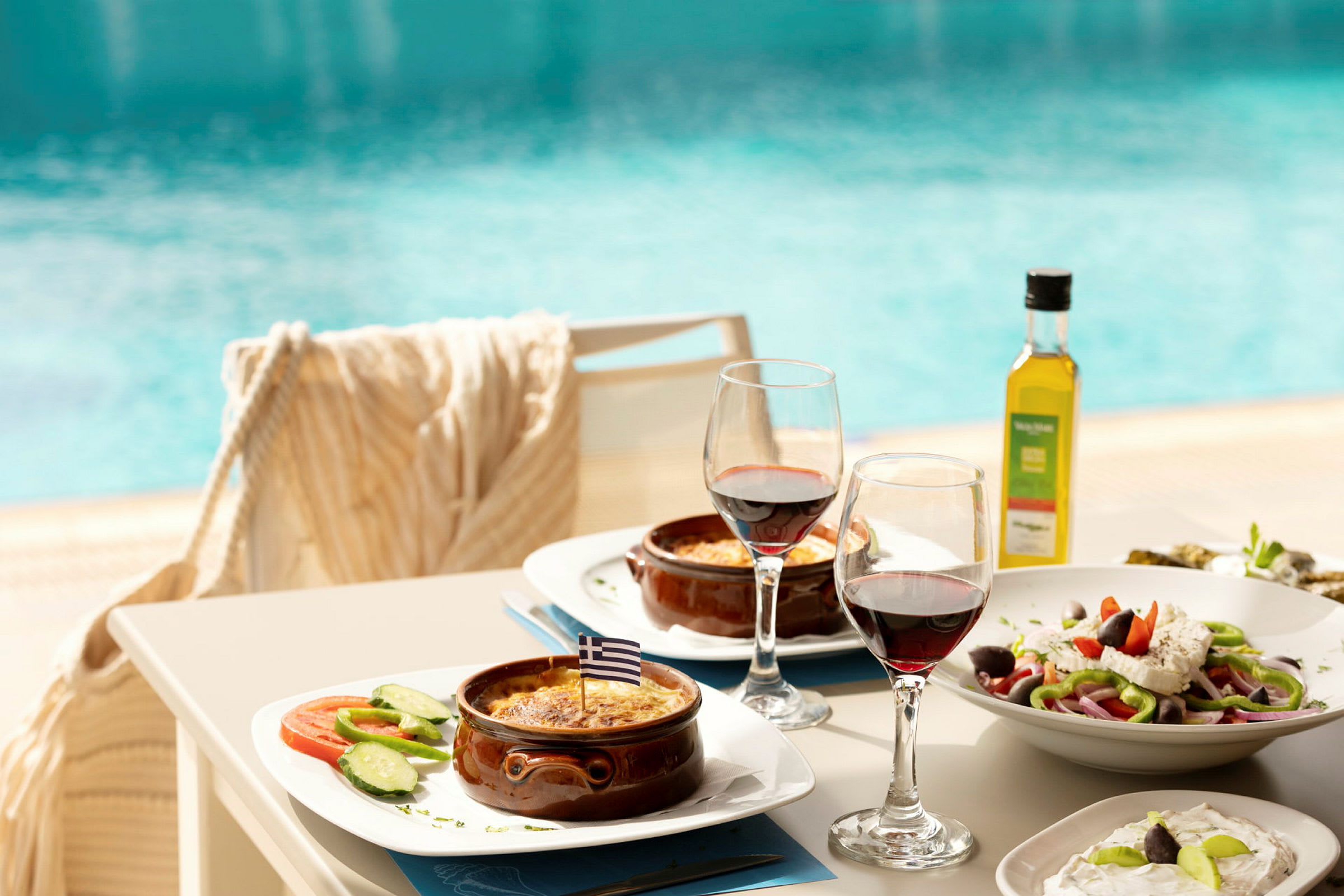 Tallrikar med grekiska rätter, två glas rött vin och en pool i bakgrunden. (AI-genererad text)