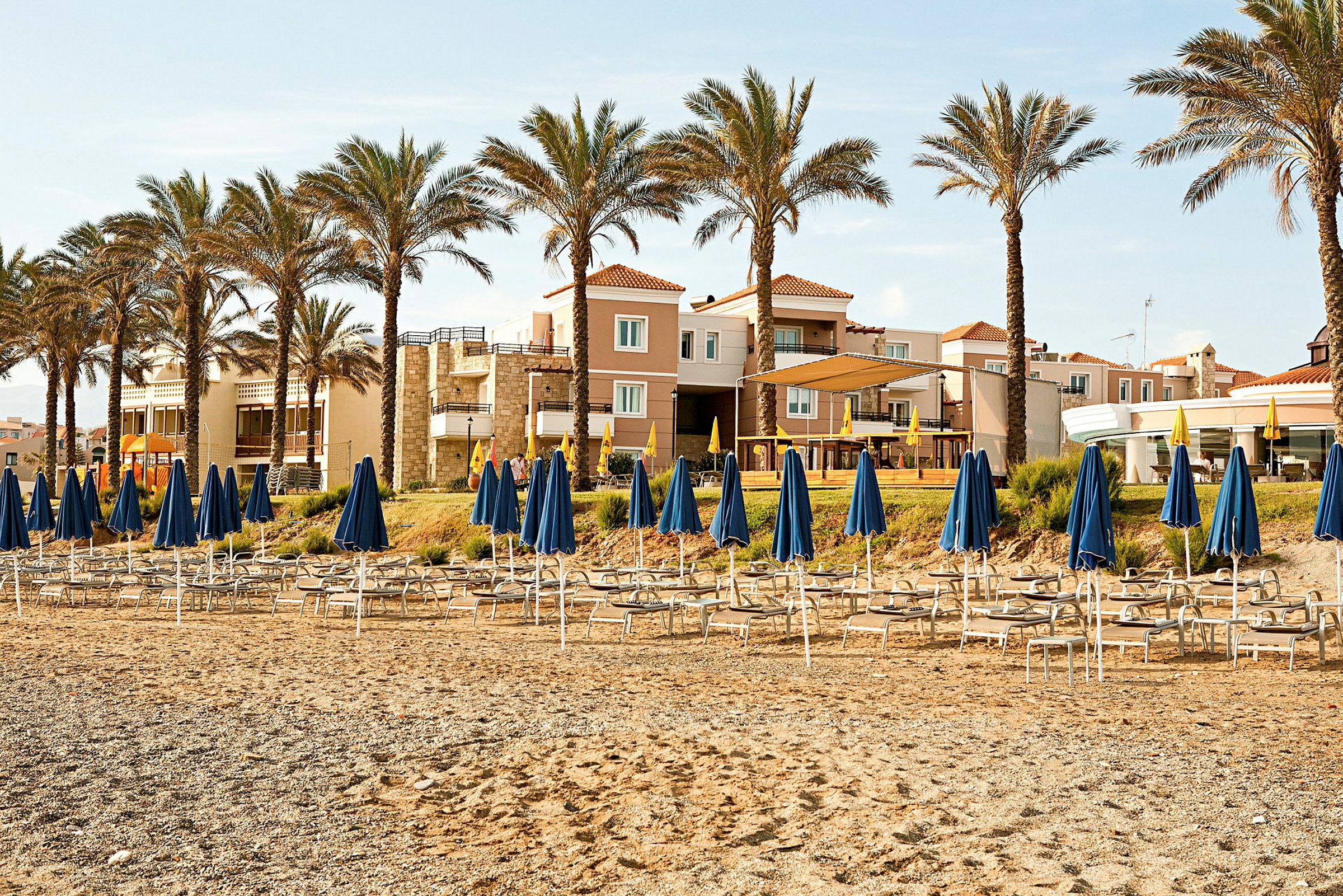 Strand med solstolar och blåa parasoller framför hotellbyggnader och palmer. (AI-genererad text)