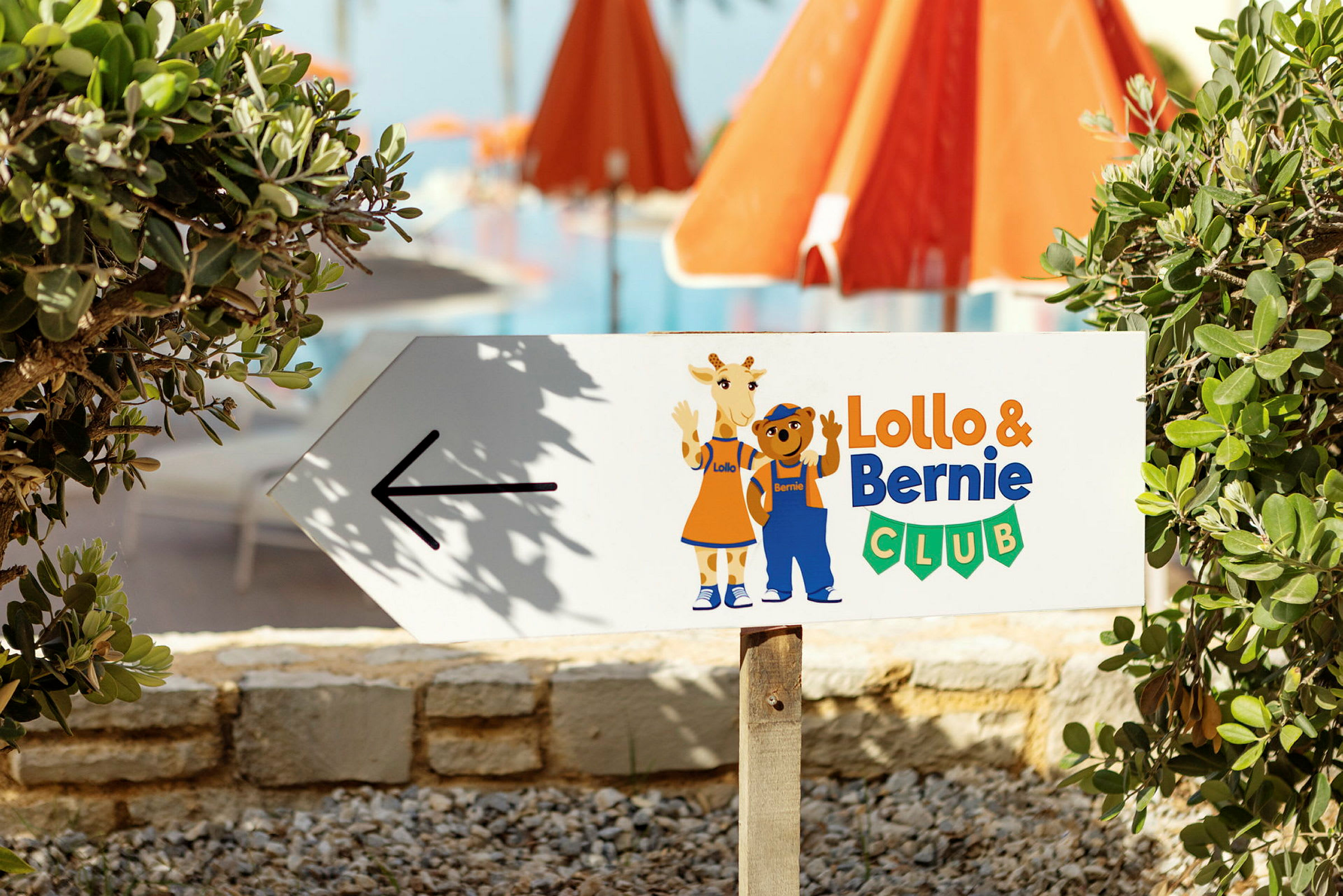 Skylt med pil och texten 'Lollo & Bernie Club', med illustrationer av Lollo och Bernie. Bakgrund med grönska och röda parasoller. (AI-genererad text)