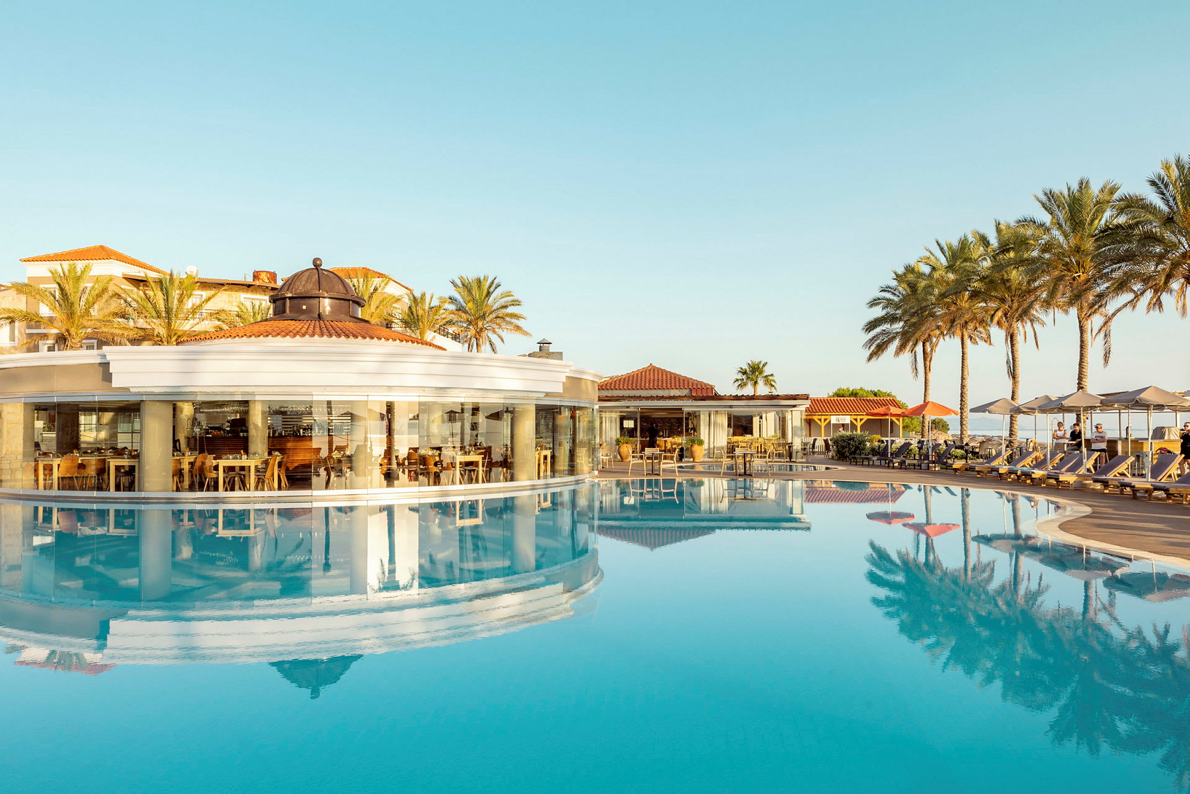 En restaurang med panoramaglas väggar vid en pool, omgiven av palmer. (AI-genererad text)