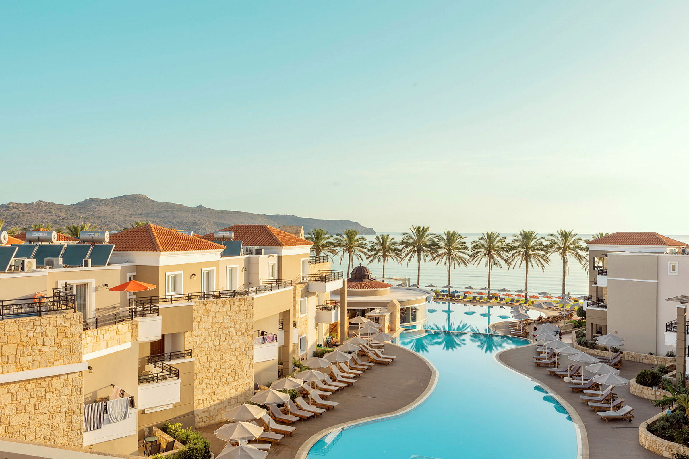 Översikt av en resort med pool och solstolar, med havsutsikt och palmer i bakgrunden. (AI-genererad text)