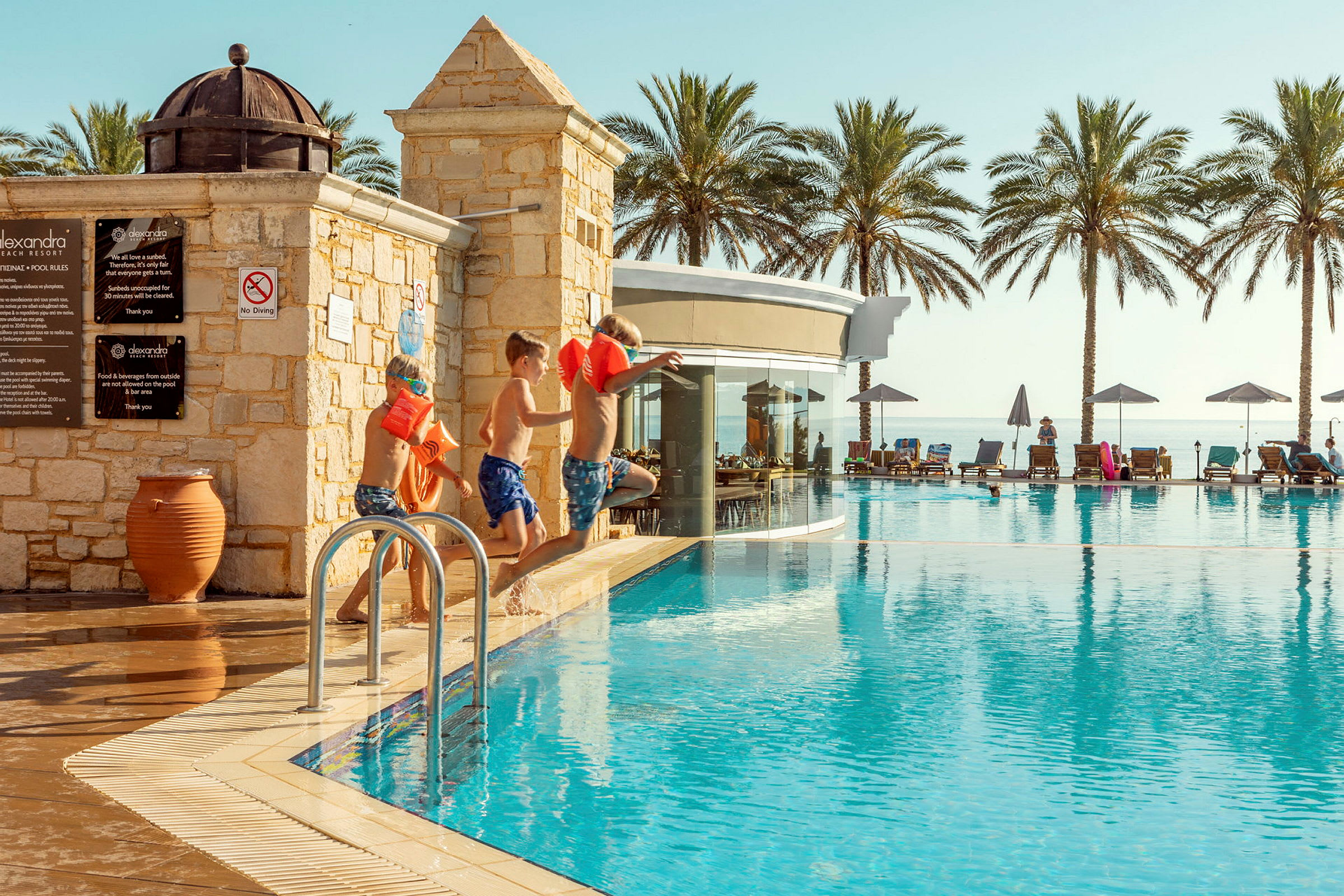 Barn hoppar i en pool i en hotellmiljö med palmer i bakgrunden. (AI-genererad text)