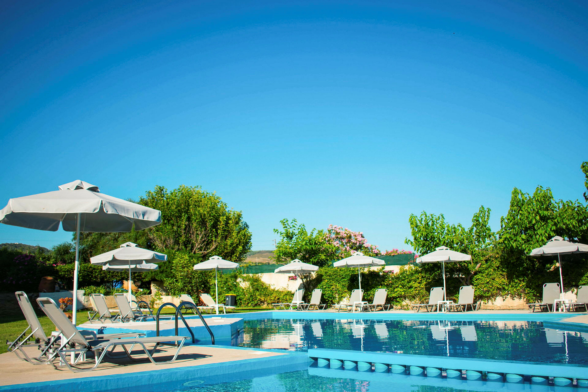 En pool med solstolar, parasoller och grönskande omgivningar under en klarblå himmel. (AI-genererad text)