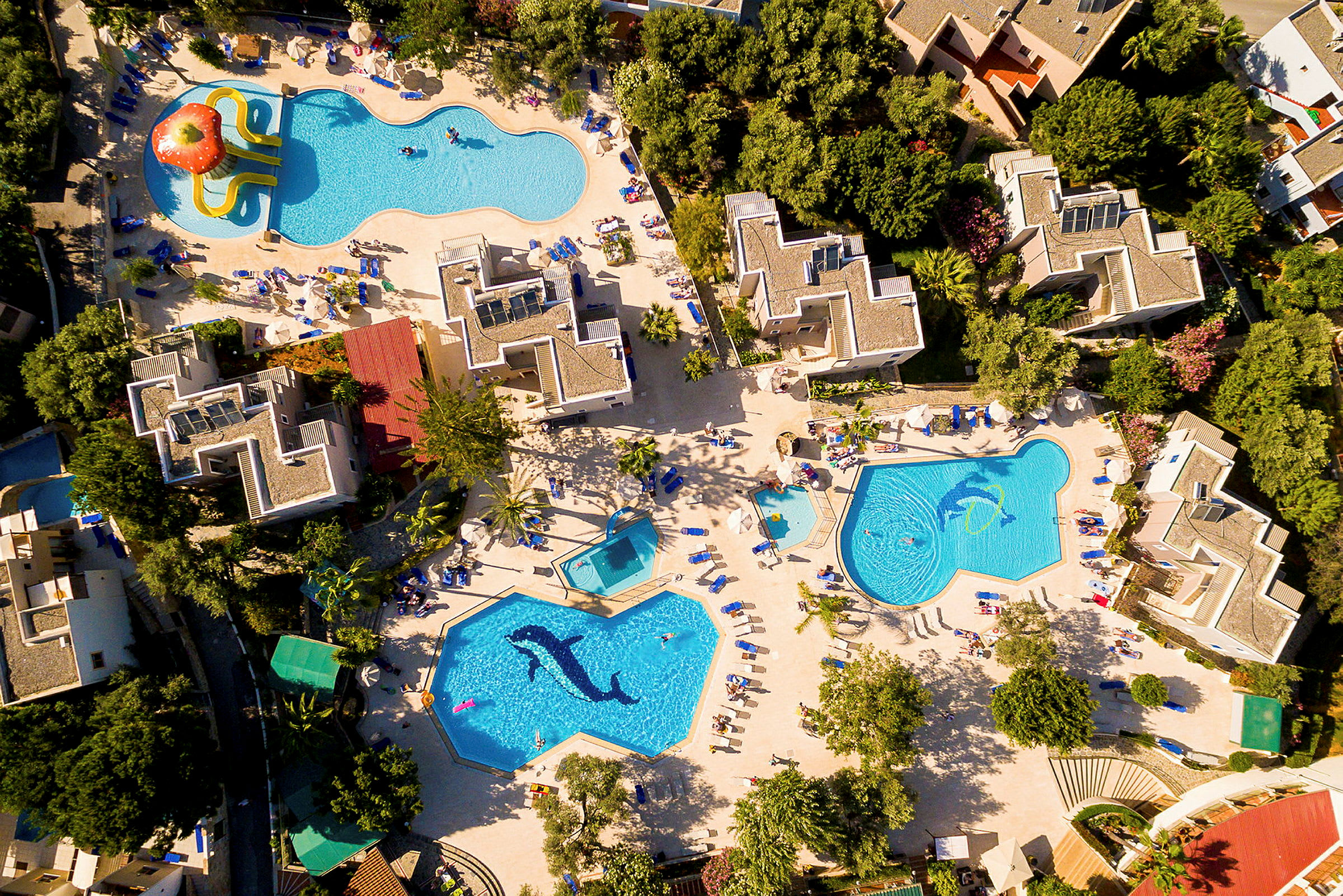 Flygfoto över ett hotellområde med flera pooler, omgivna av solstolar och grönskande trädgårdar. (AI-genererad text)