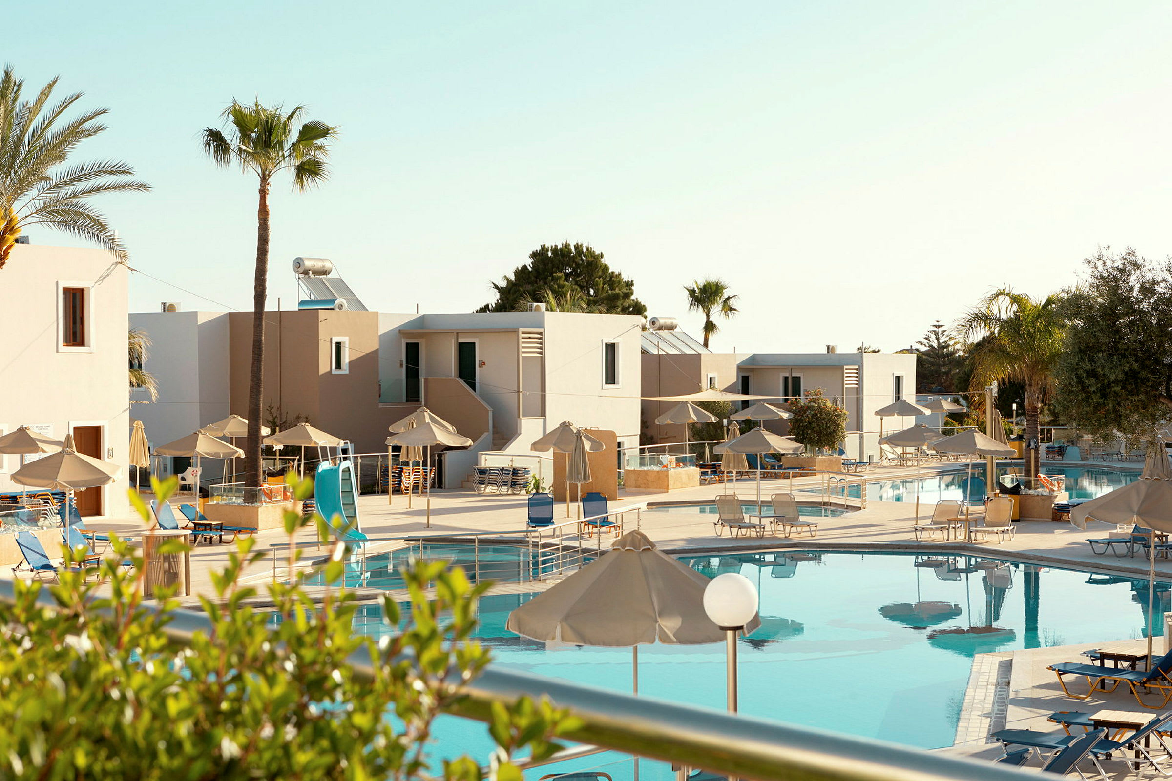 Bild på en hotellpool med solstolar och parasoller, omgiven av palmer och byggnader. (AI-genererad text)