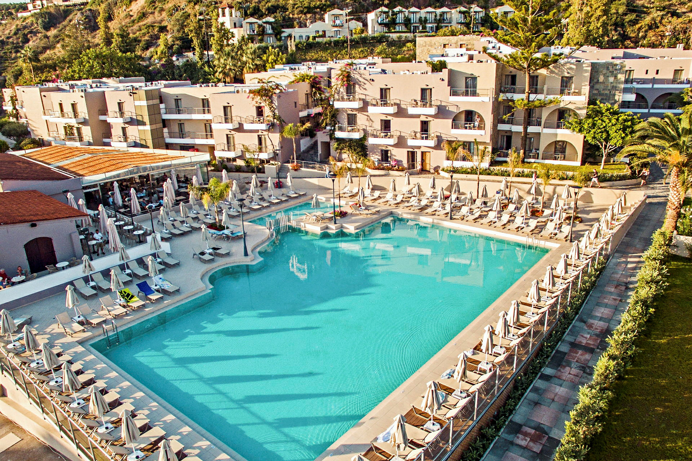 En stor blå swimmingpool omgiven av solstolar och parasoller framför en hotellbyggnad. (AI-genererad text)