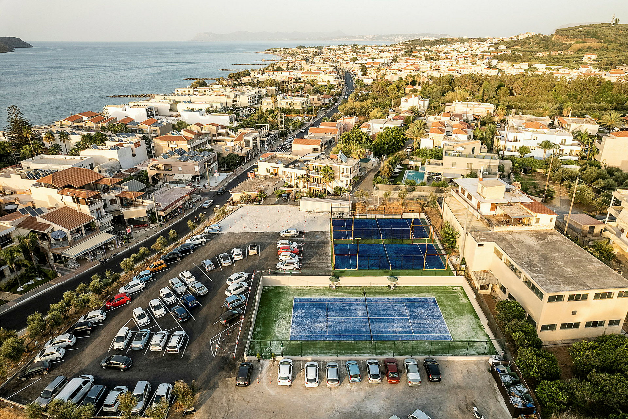 Flygfoto över en stad vid kusten, där man ser tennisbanor, parkerade bilar och byggnader. (AI-genererad text)