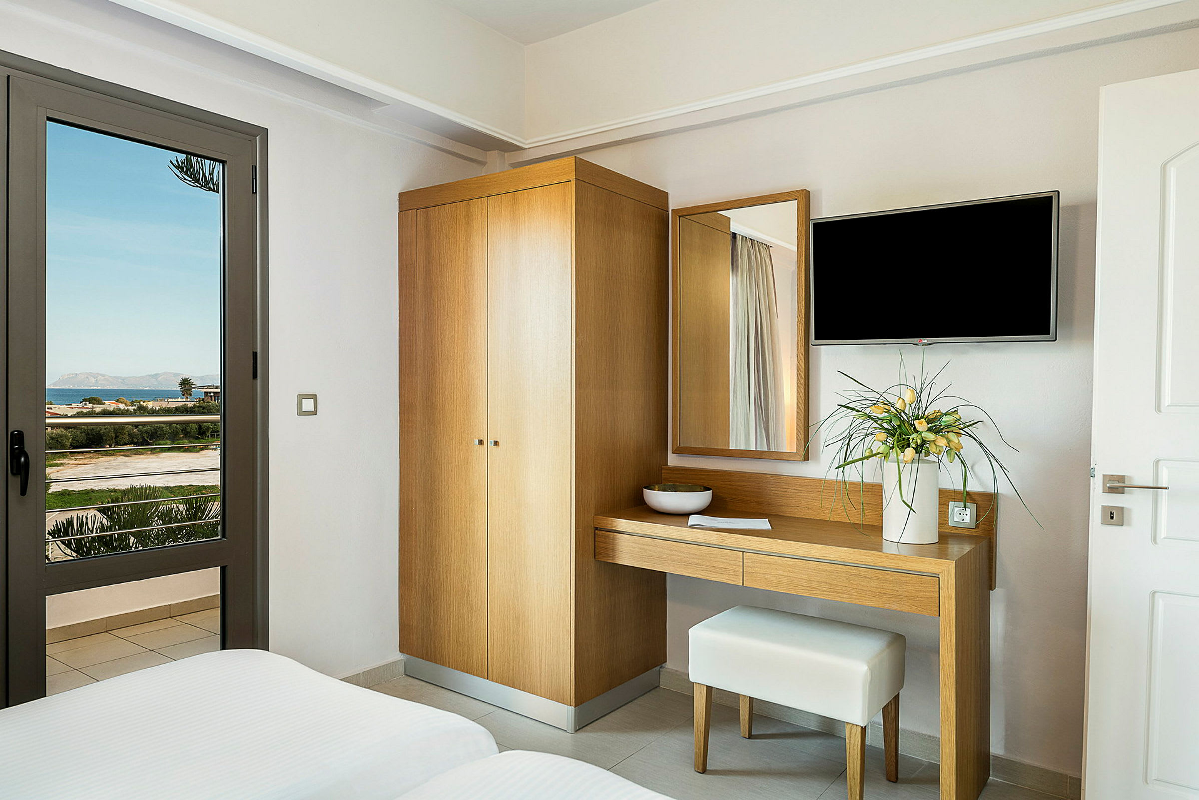Hotellrum med garderob, skrivbord och blomdekoration, samt utsikt över landskap genom glasdörr. (AI-genererad text)