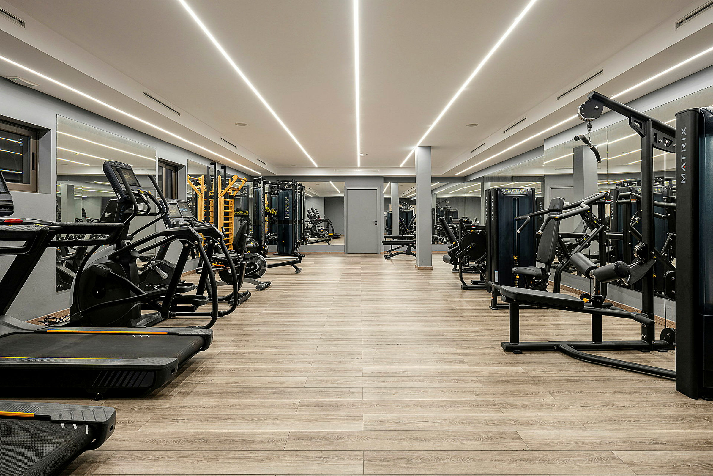 Ett modernt gym med träningsutrustning, inklusive löpband och styrketräningsmaskiner, med trägolv och speglar längs väggarna. (AI-genererad text)